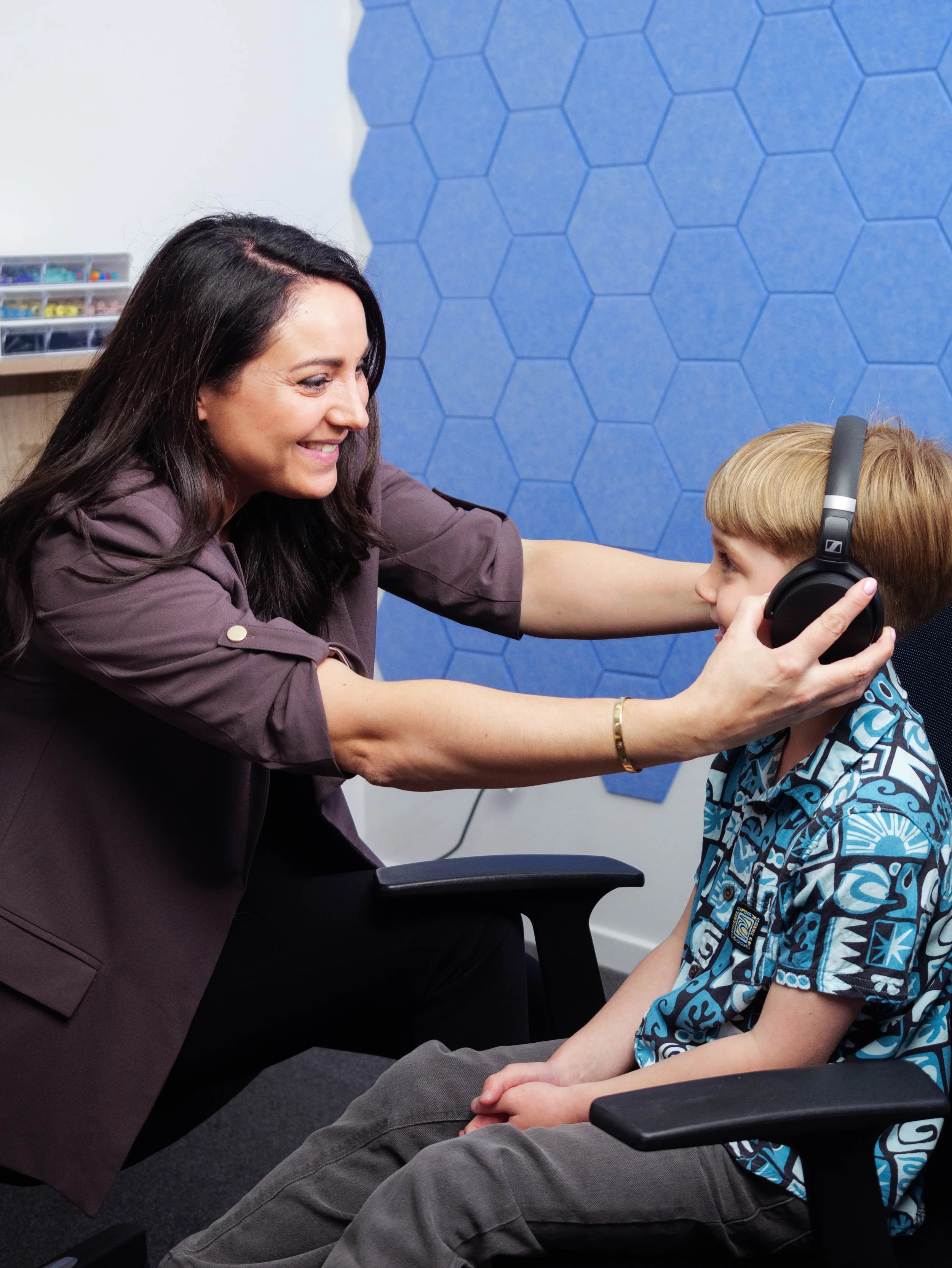Katerin Hobson is helping a boy put on headphones in a room with blue hexagon-patterned wall. Testing his hearing.