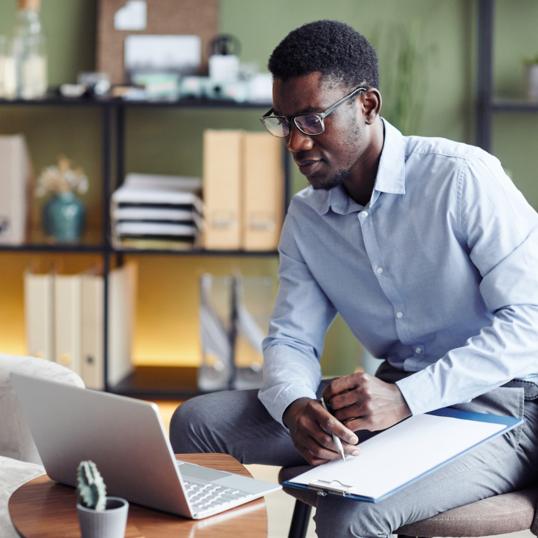 Man writing in a notebook while reviewing his laptop, representing online burnout therapy for professionals