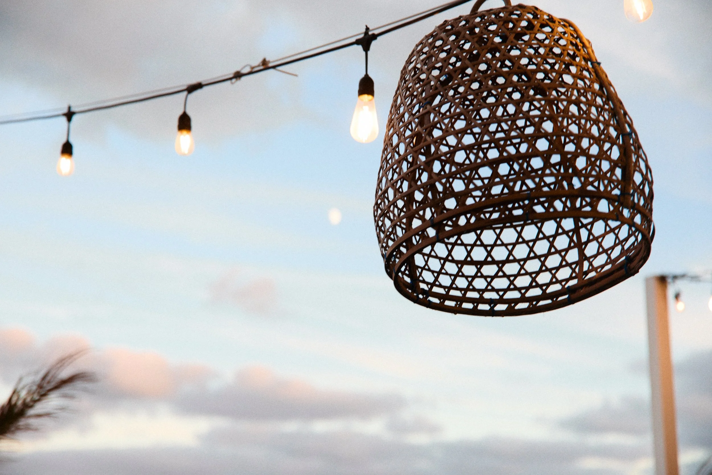 Outdoor string lights with bulbs hanging above a wicker lampshade against a cloudy sky