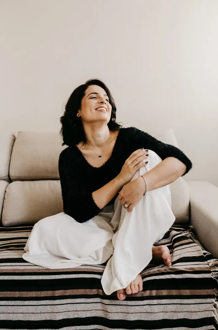 A woman sitting on a sofa, smiling and laughing, wearing a black sweater and white pants, with a neutral background.