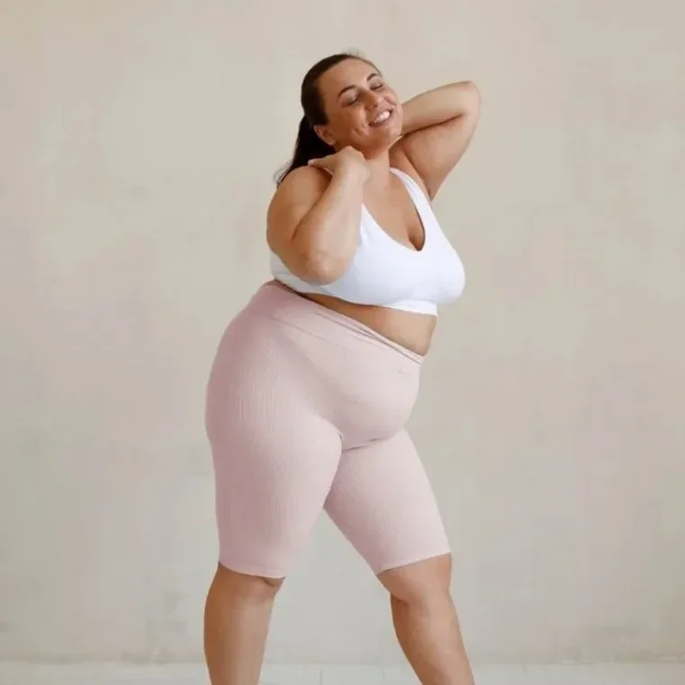 A woman smiling with eyes closed, wearing a white sports bra and pink shorts, standing against a plain beige background.