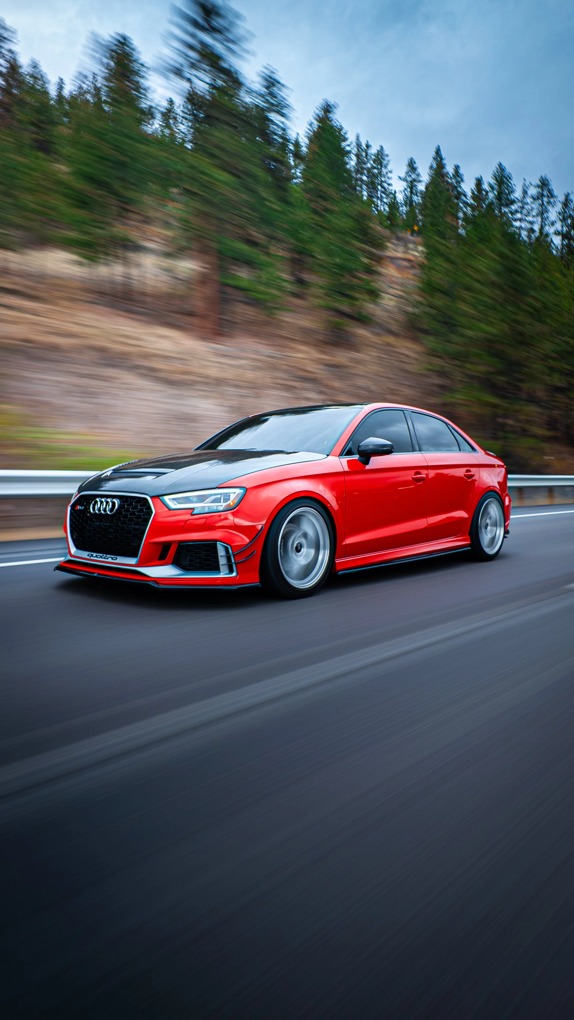 Red sports car with black hood traveling on a highway with a blurred background of trees and mountains.