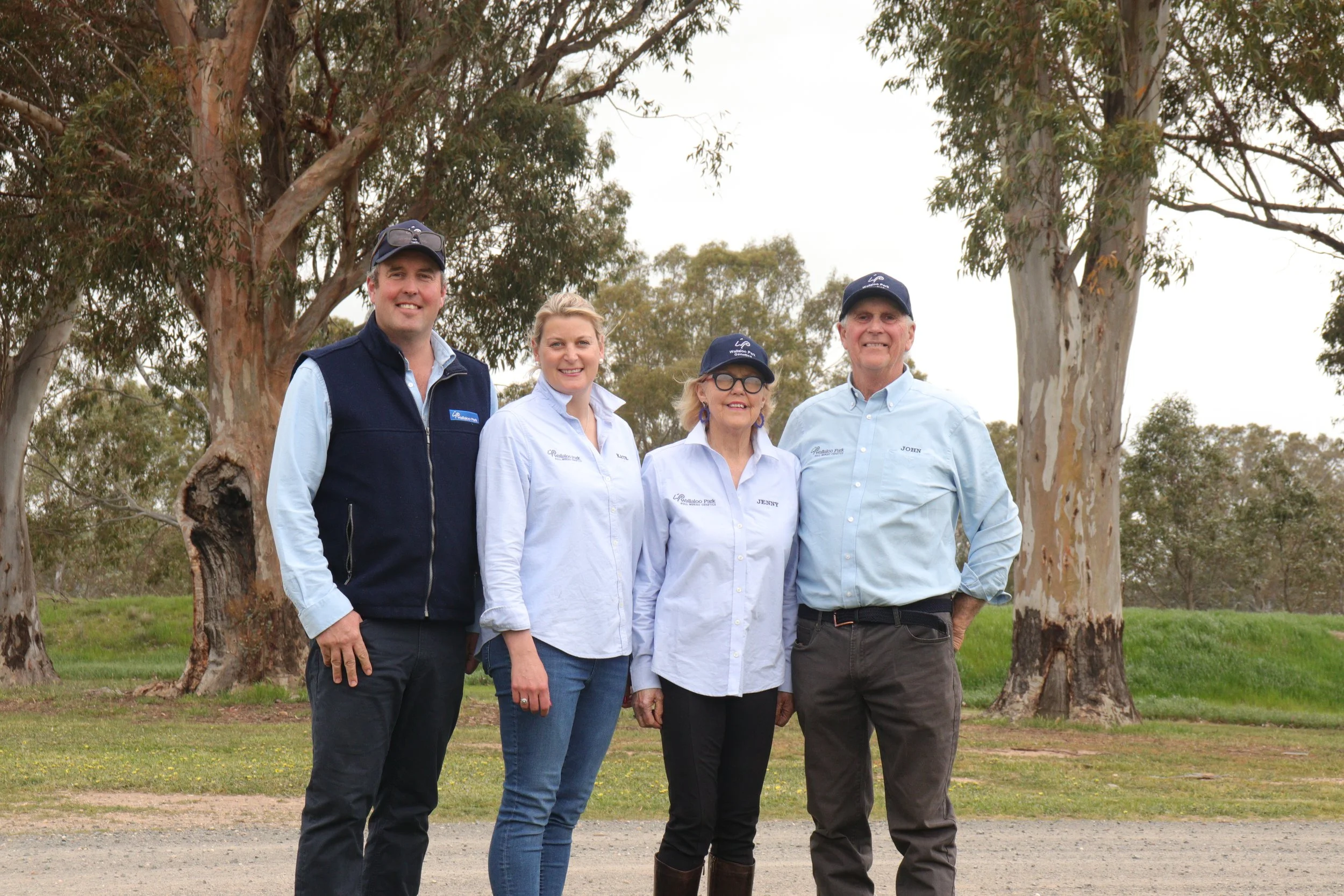 Four people standing outdoors in front of trees, smiling and posing for a photo. They are dressed in casual and work attire.