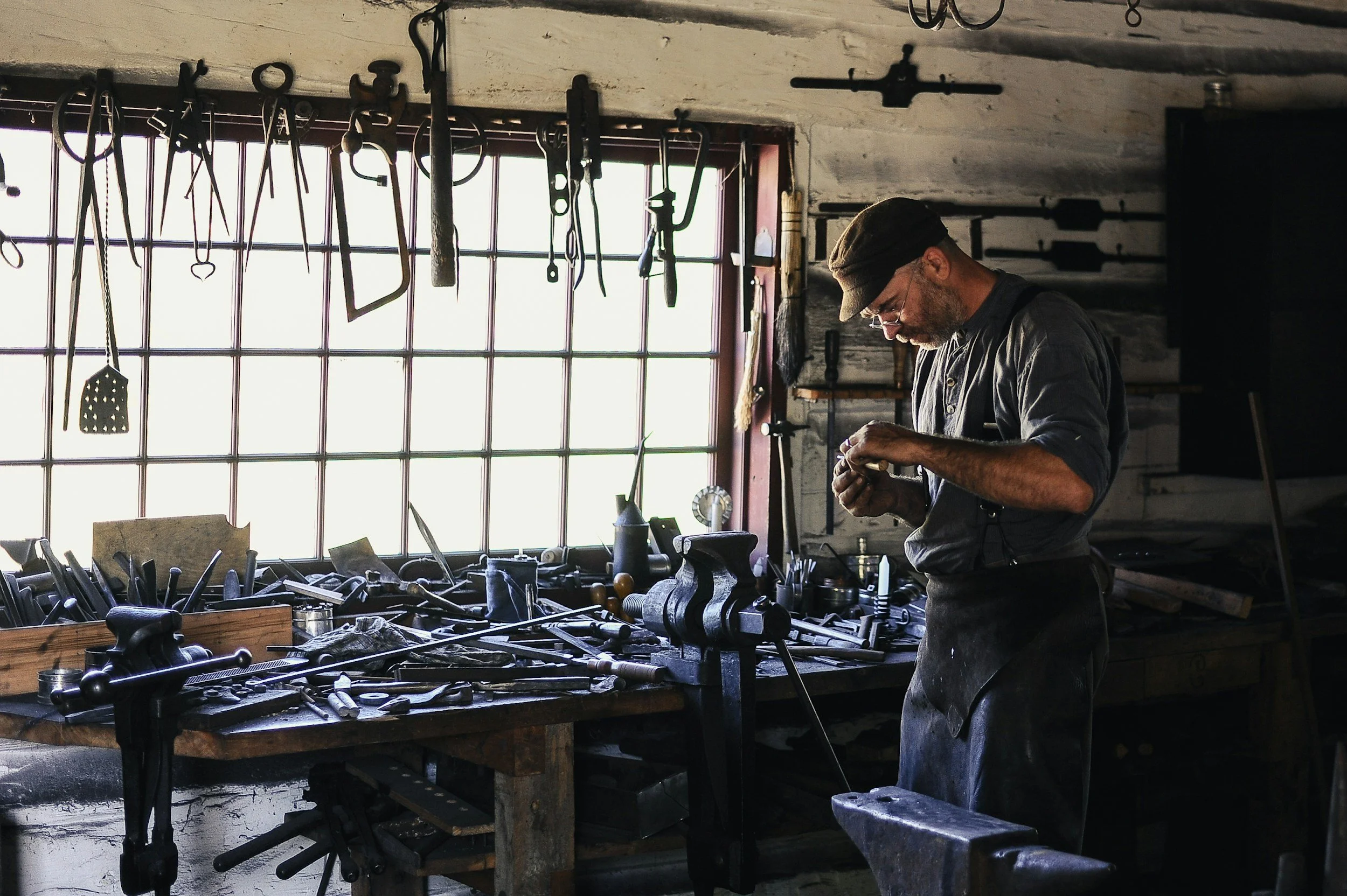 A man working in a metalworking shop. He is wearing a cap and apron, standing at a cluttered workbench filled with tools and metal pieces. The shop has a large window with sunlight streaming through, and various tools hanging from the ceiling.