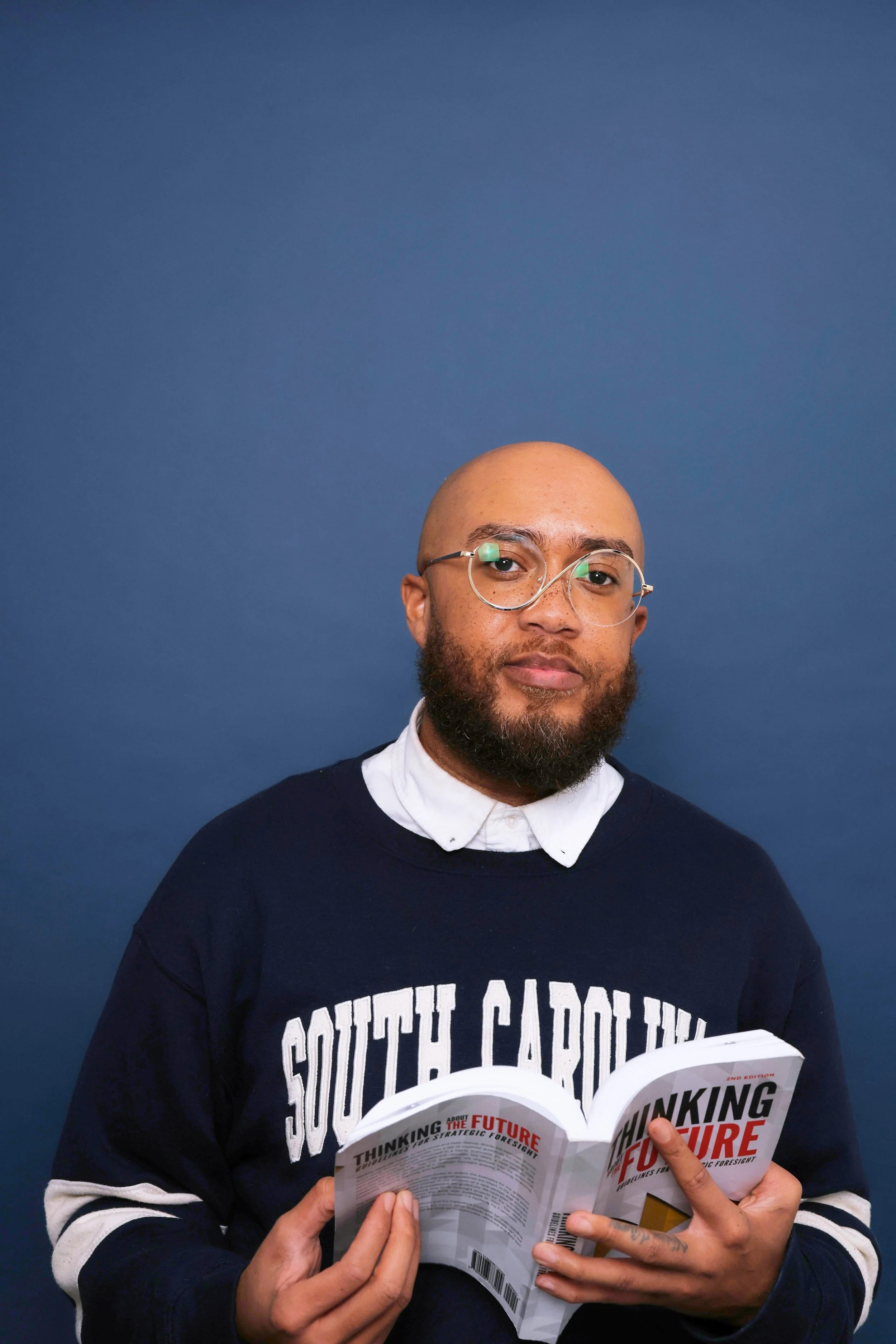 A man with a bald head, beard, and glasses, wearing a dark sweatshirt with white lettering, reads a book titled 'Thinking About the Future' in front of a blue background.