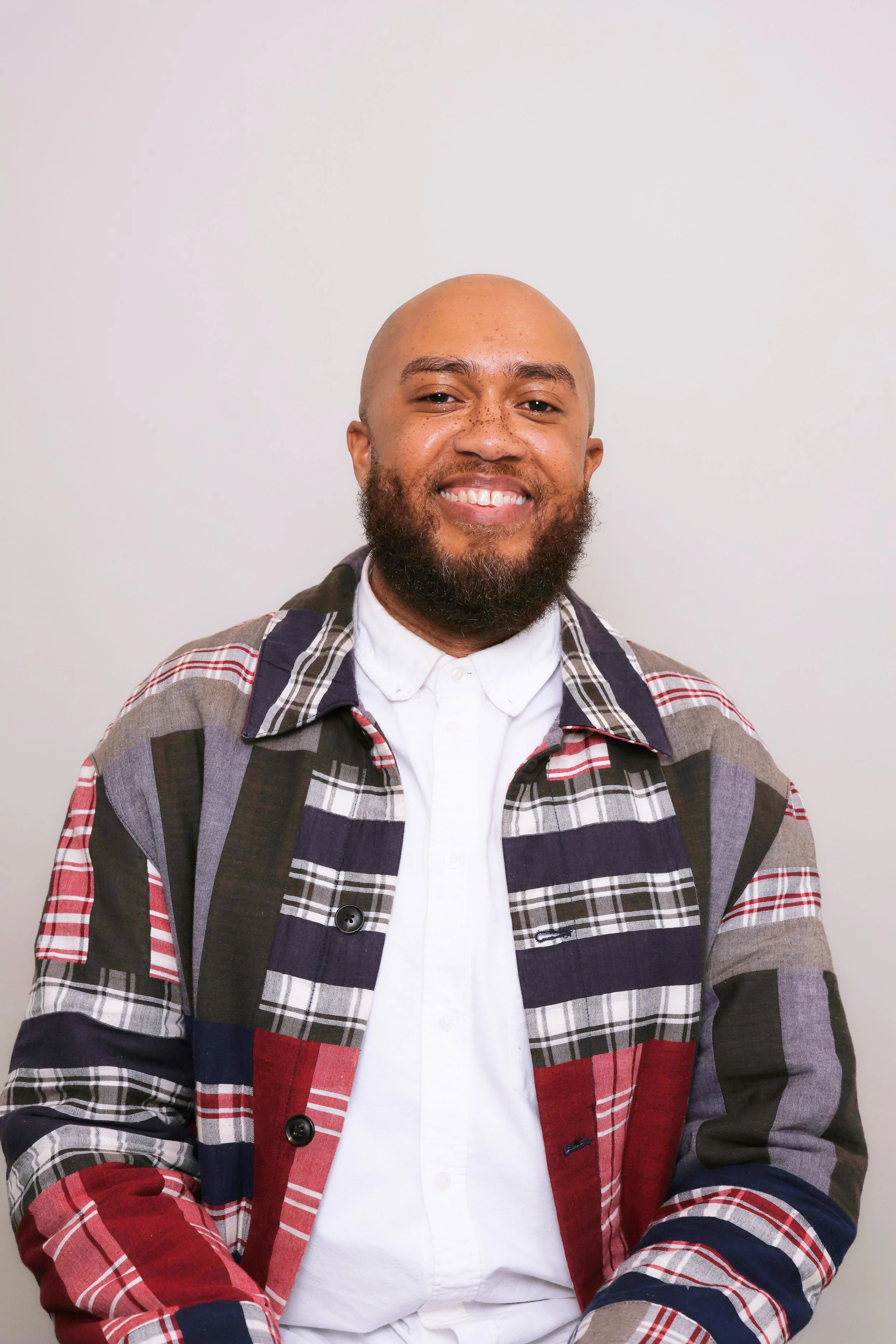 A smiling man with a beard and a bald head, wearing a plaid jacket over a white shirt, standing against a plain white background.