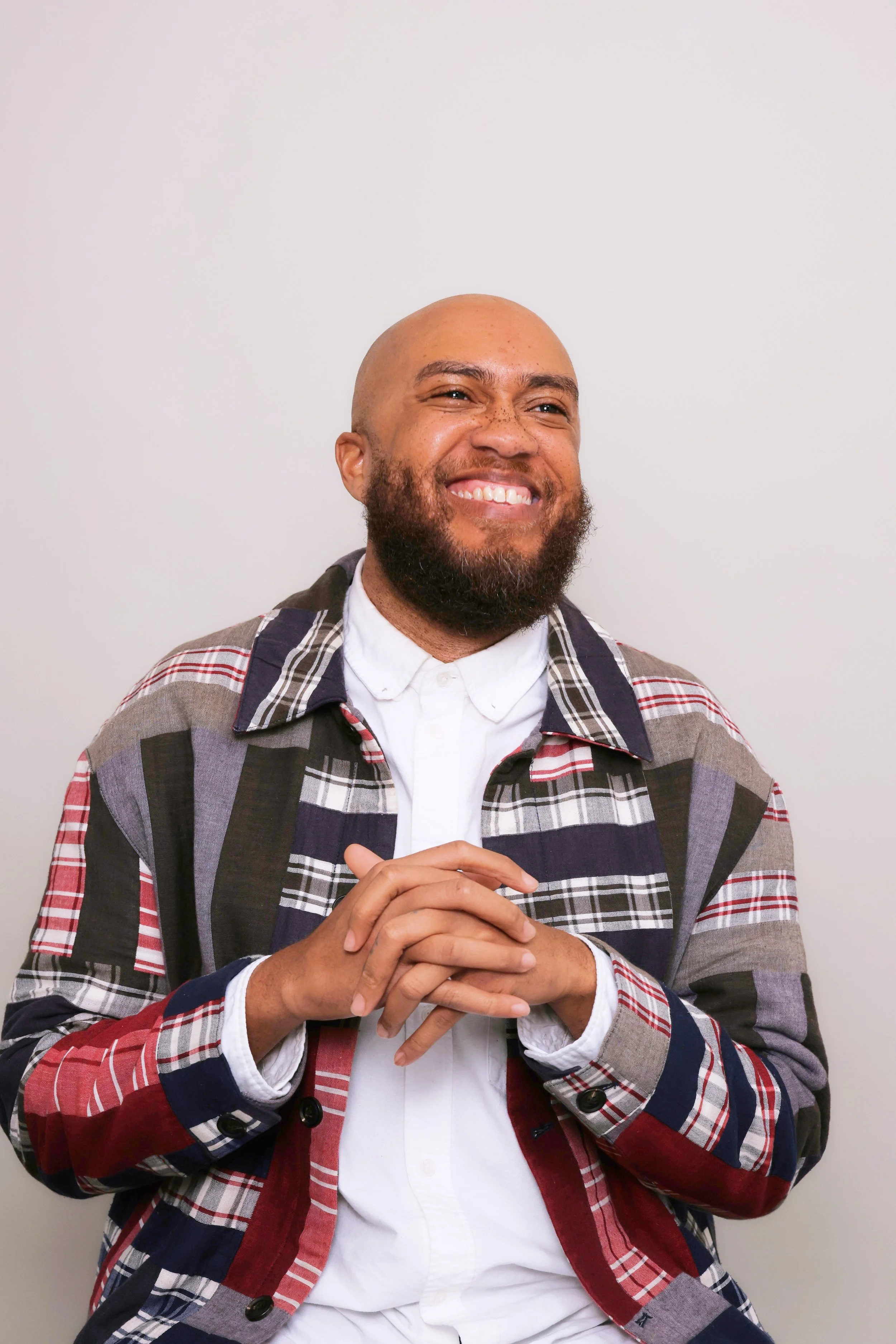 A man with a big beard and mustache, smiling with teeth showing, wearing a white shirt and a multicolored plaid jacket, standing against a plain white wall.