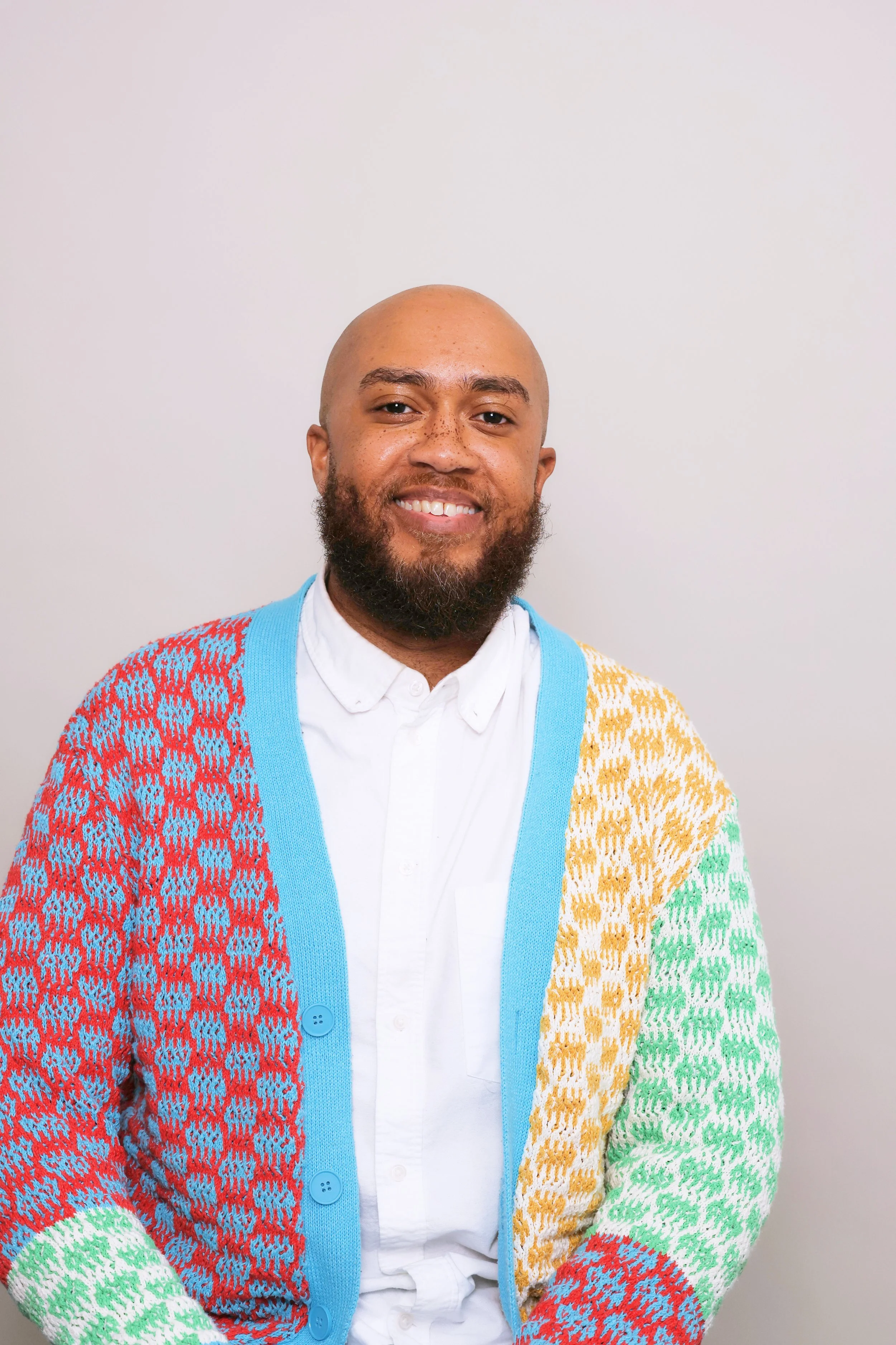 A smiling man with a beard and a shaved head, wearing a colorful patterned cardigan, a white button-up shirt, and standing against a plain light-colored background.