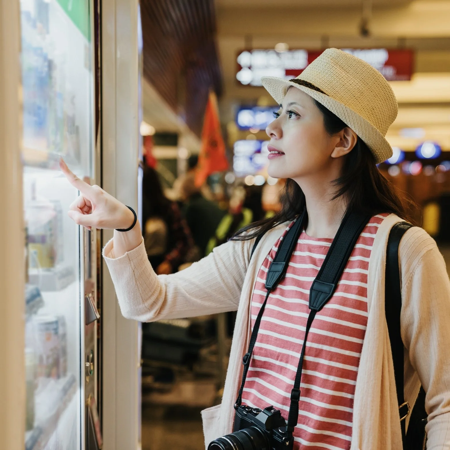 A woman with a camera around her neck wearing a striped shirt, beige cardigan, and straw hat, selects an item from a vending machine in an indoor setting like an airport or train station.
