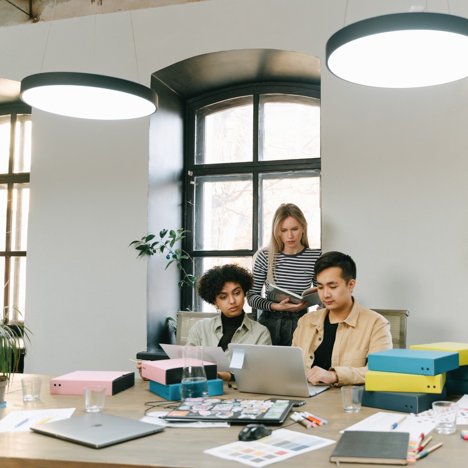 Three young people working together at a cluttered table in a modern office with large windows and hanging lights.
