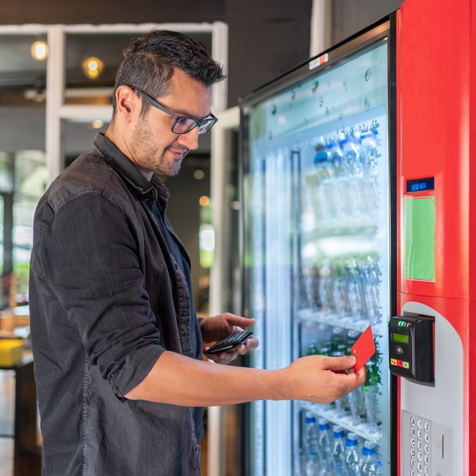 A man in a black shirt and glasses using a vending machine, holding a red card in his hand.