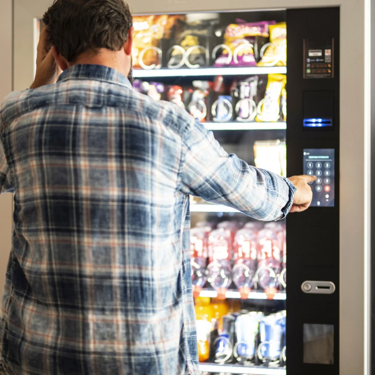 A man in a plaid shirt using a vending machine with snacks on the shelves.