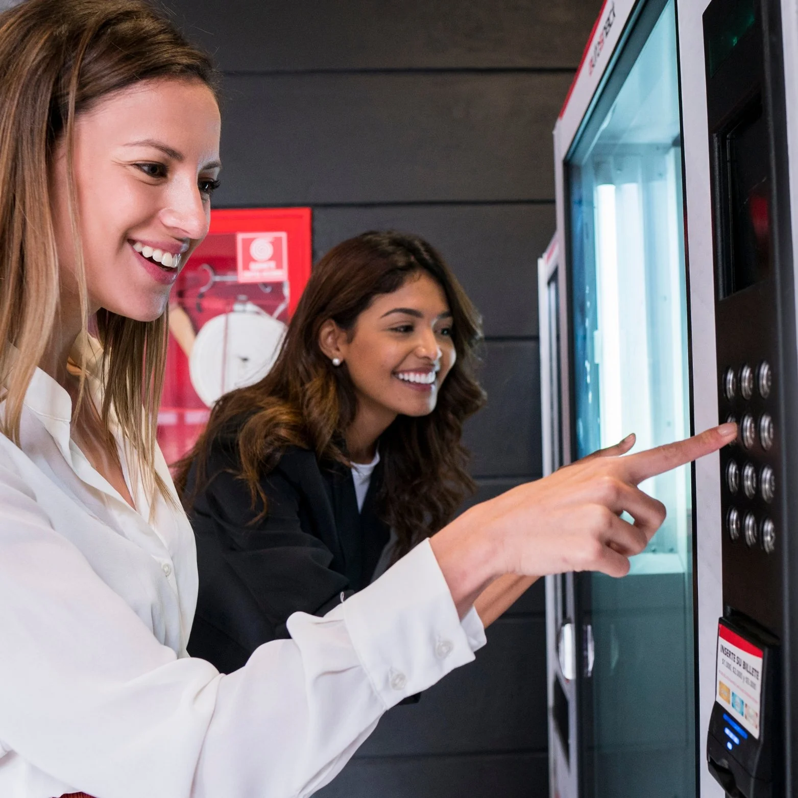 Two women smiling and selecting a beverage from a vending machine.