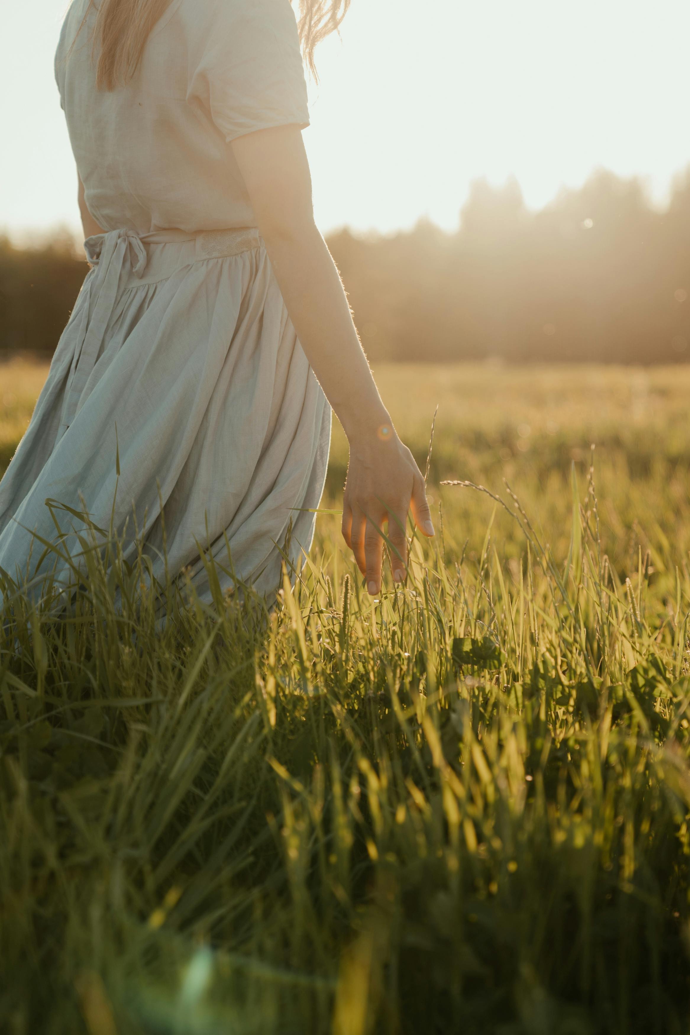 A person with light-colored dress and long hair touching grass in a field during sunset.