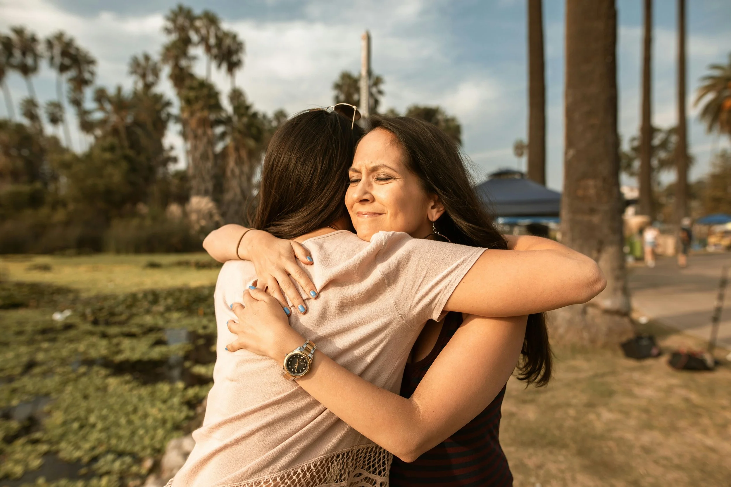 Two women hugging outdoors with palm trees, blue sky, and a few people in the background.
