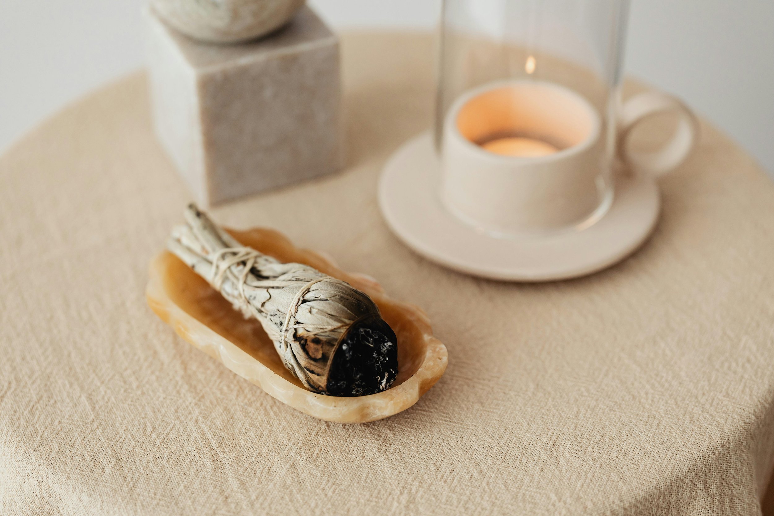 A smudge stick of palo santo wood placed on a shell, with a lit candle in a glass holder and a beige textured tablecloth.
