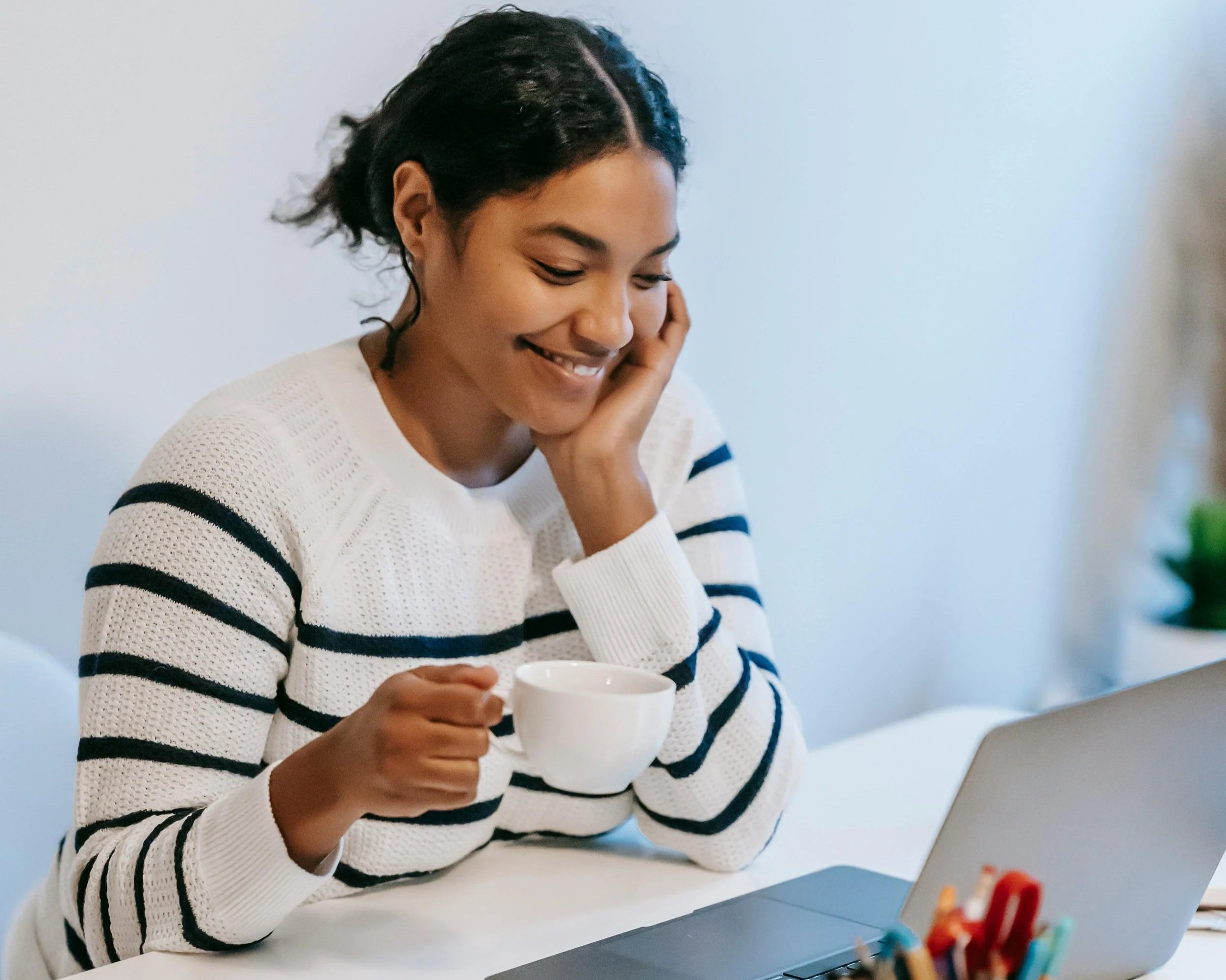 A woman with short curly hair smiling and holding a white mug, sitting at a table with a laptop in front of her.