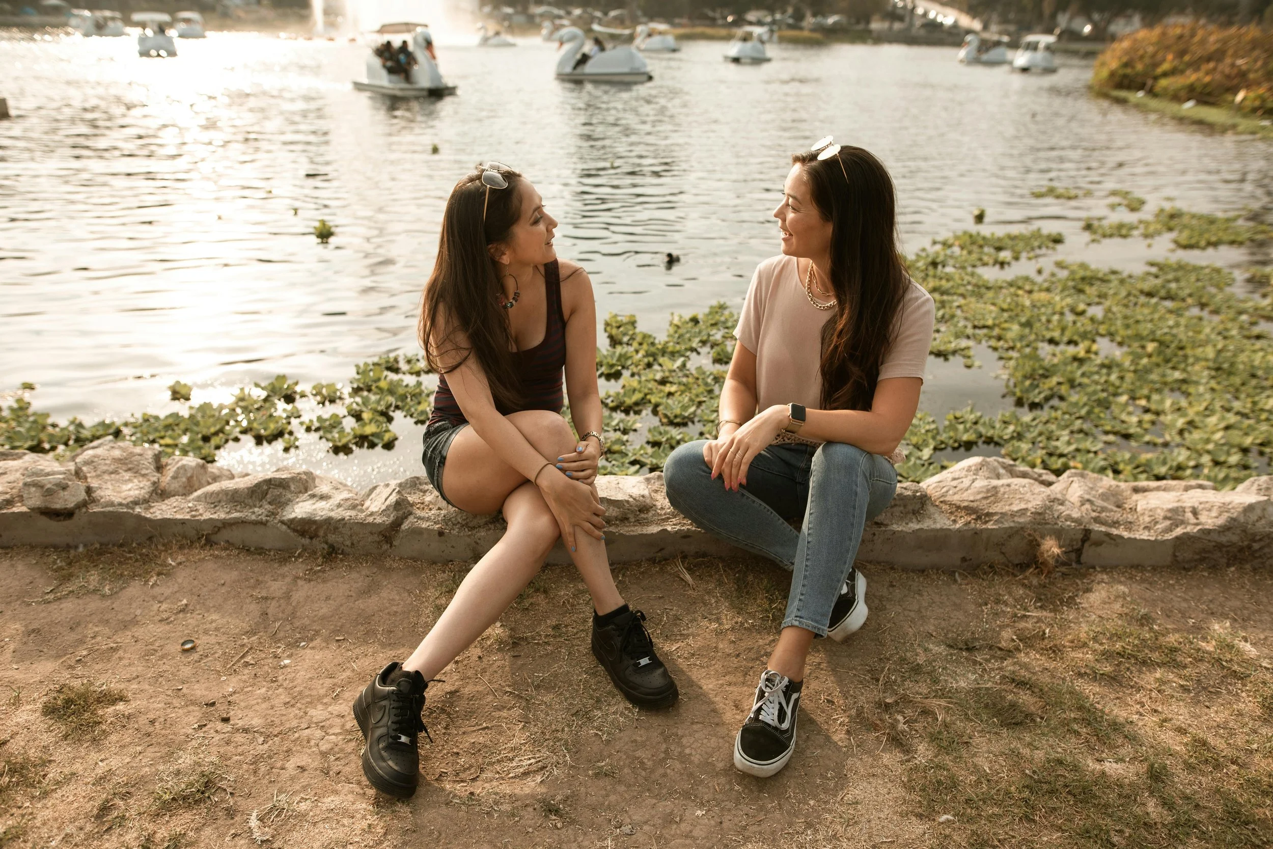 Two women sitting on a stone border by a lake, engaged in conversation, with pedal boats shaped like swans on the water in the background.