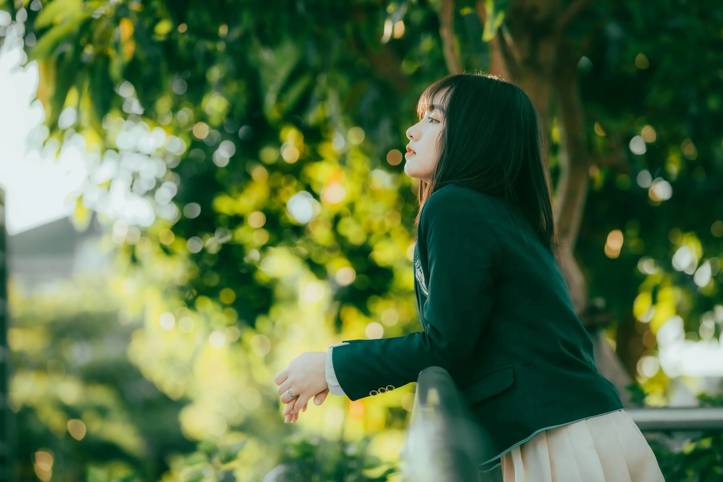 A young woman in a dark blazer leaning on a railing outdoors amidst green trees, gazing to the side in profile.