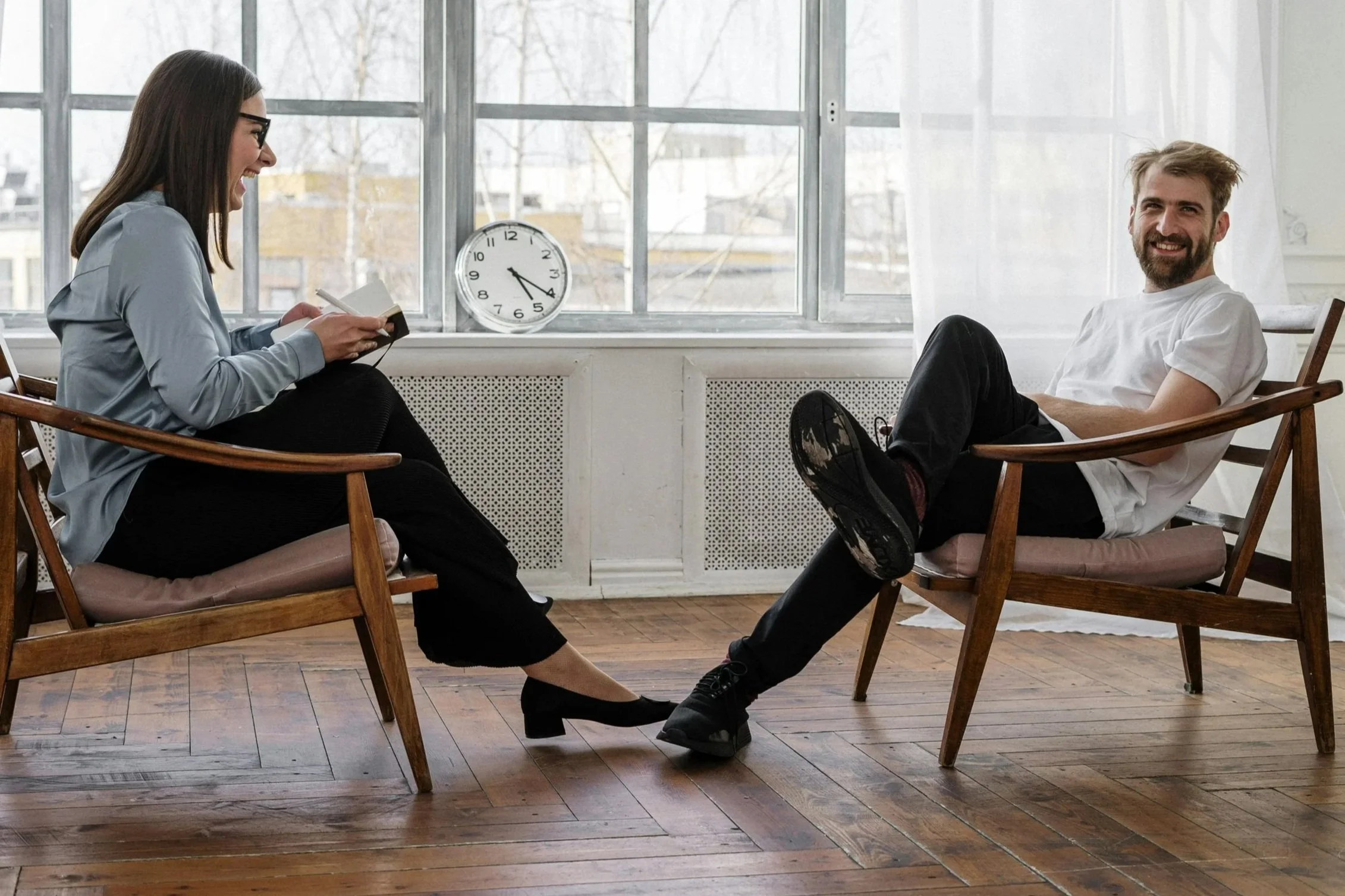 A woman and a man sitting in chairs facing each other, smiling and laughing in a bright room with large windows and wooden floors.