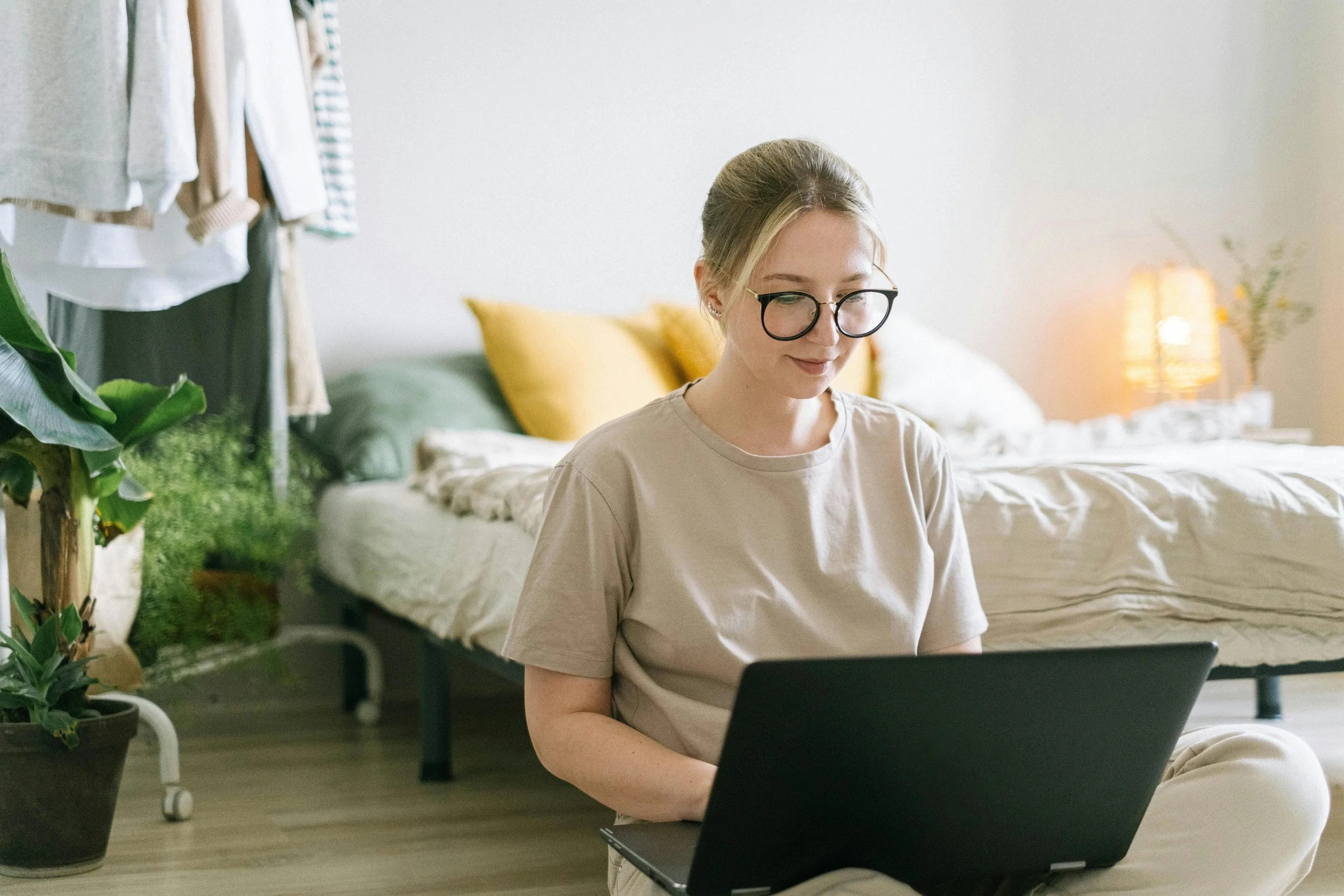 A young woman with blonde hair, glasses, and a beige t-shirt sitting on the floor and working on a laptop in a bedroom decorated with plants, a bed with mustard-colored pillows, and a lit nightstand in the background.