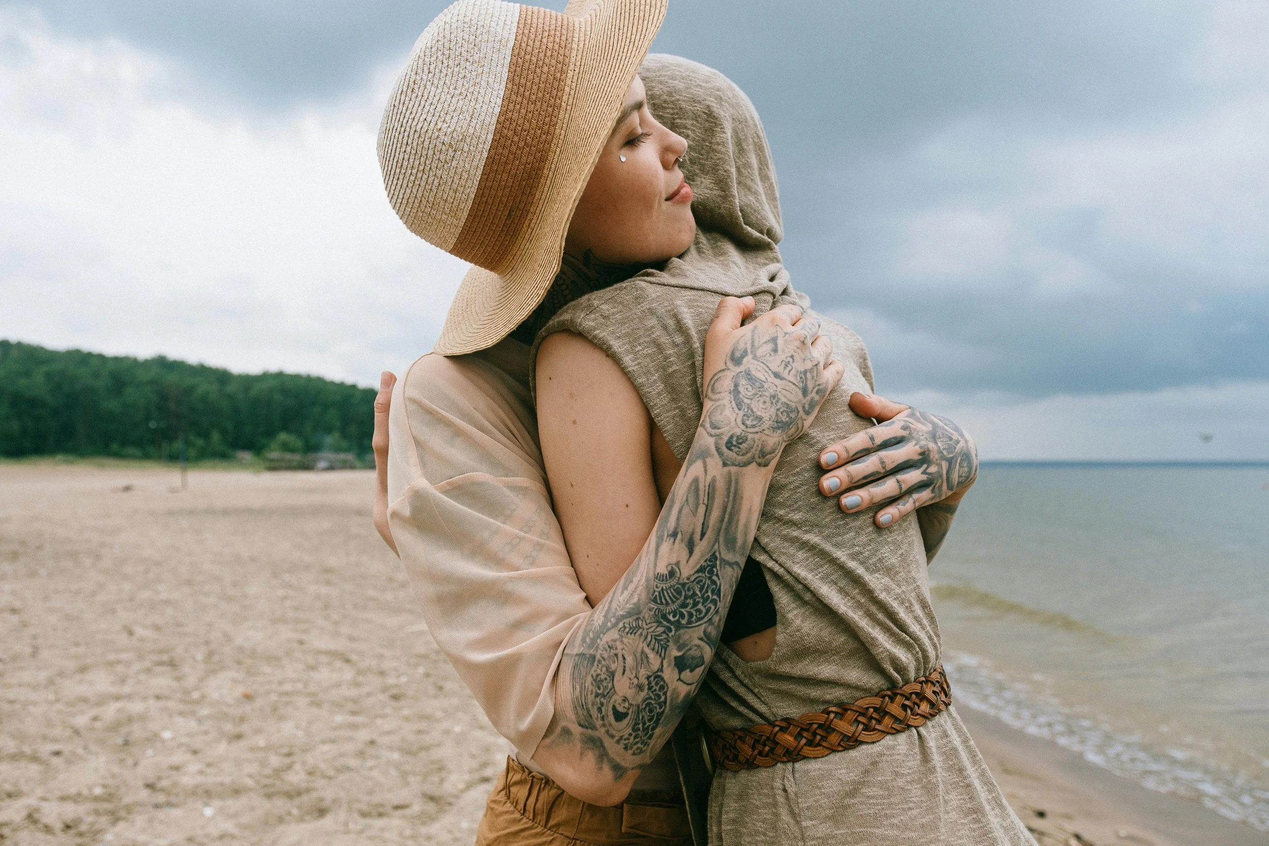 A woman with tattoos on her arm hugging a child with a hoodie on a beach, overcast sky visible in the background.
