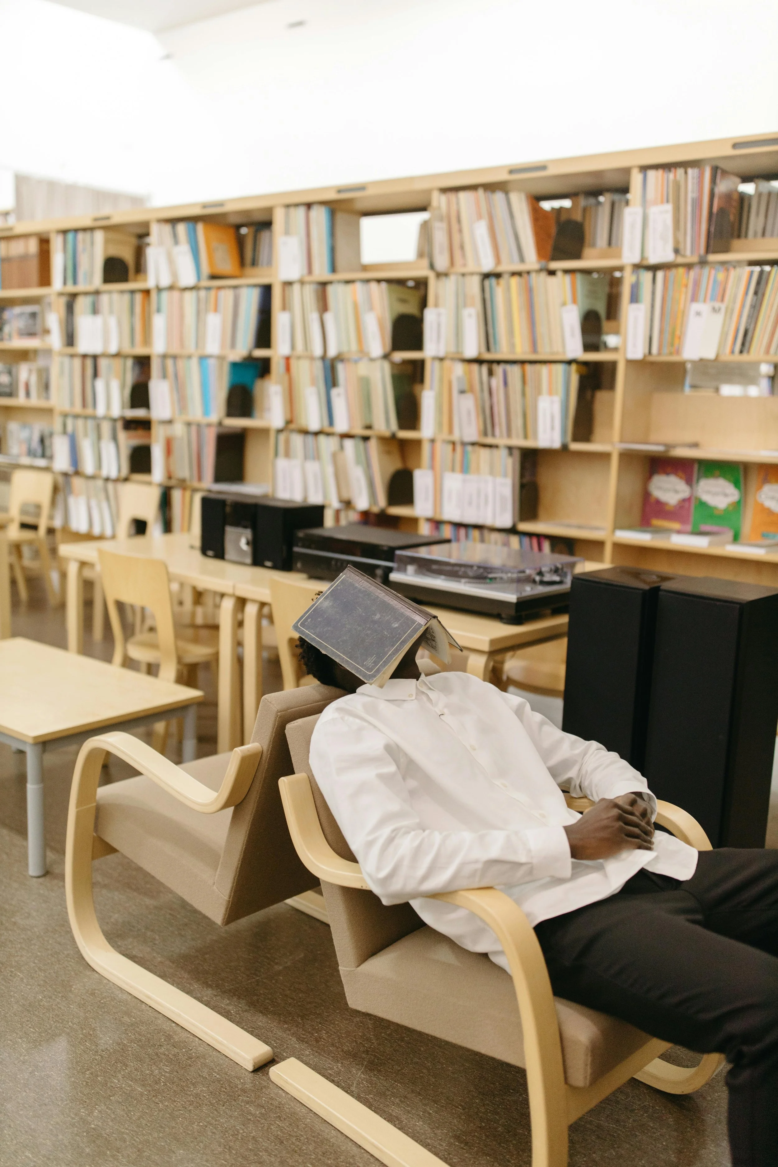 A person in a white shirt is sitting on a bench with a book on their face in a library. The library has wooden bookshelves filled with books and a table with record players and speakers.