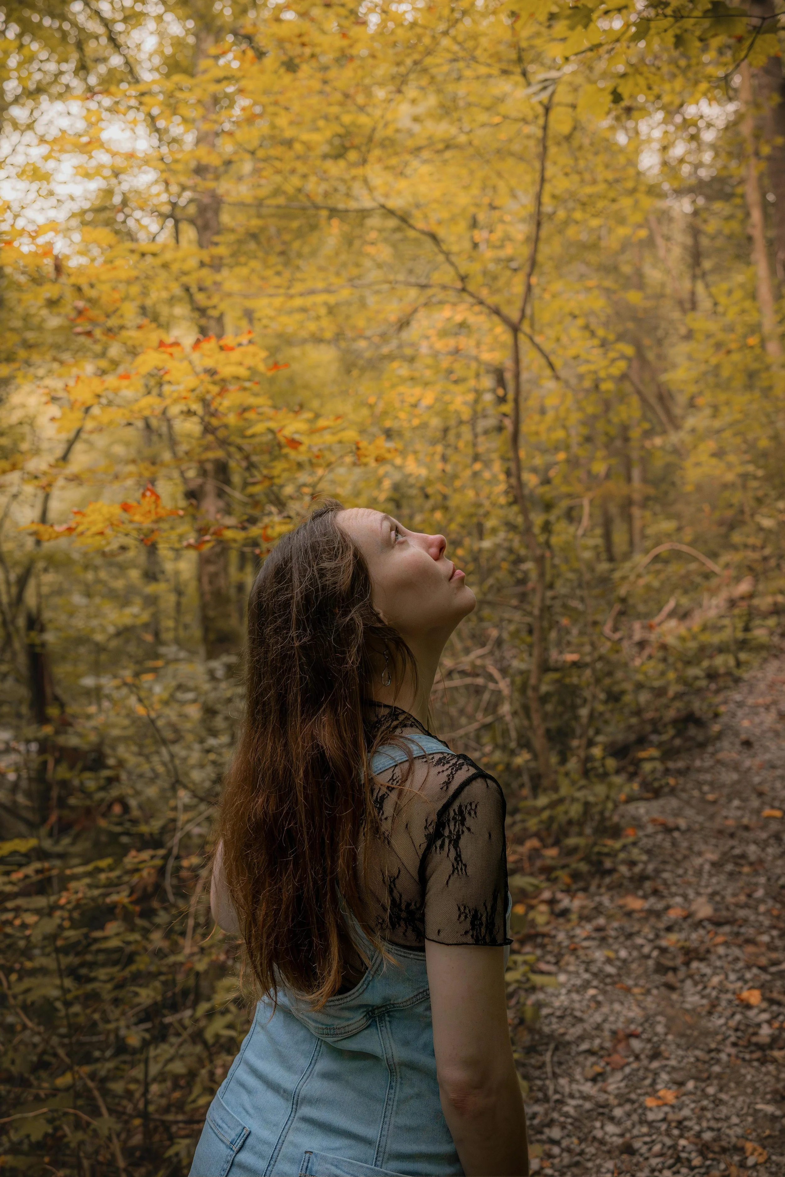 A young woman with long brown hair, wearing a black lace top and denim overalls, stands on a forest trail surrounded by autumn-colored foliage, gazing upward with a peaceful expression.