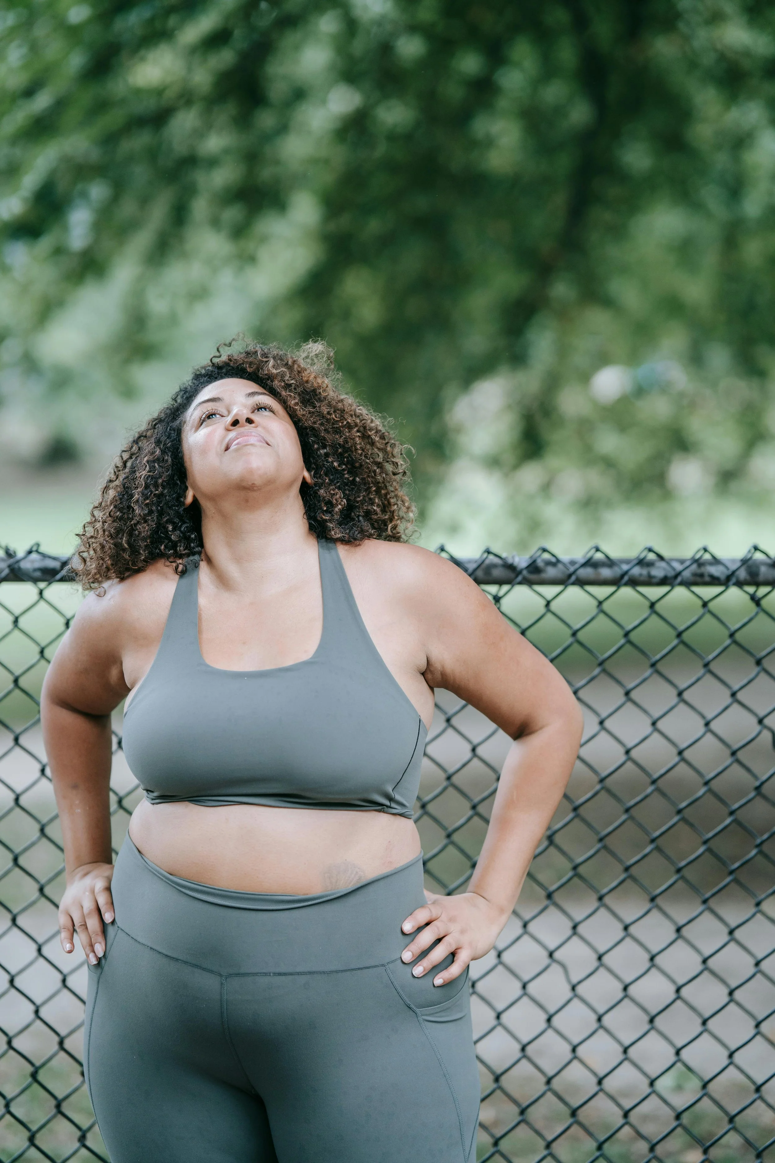 A woman in athletic wear standing outdoors with her hands on her hips, looking up with a determined expression, in front of a chain-link fence and green trees.