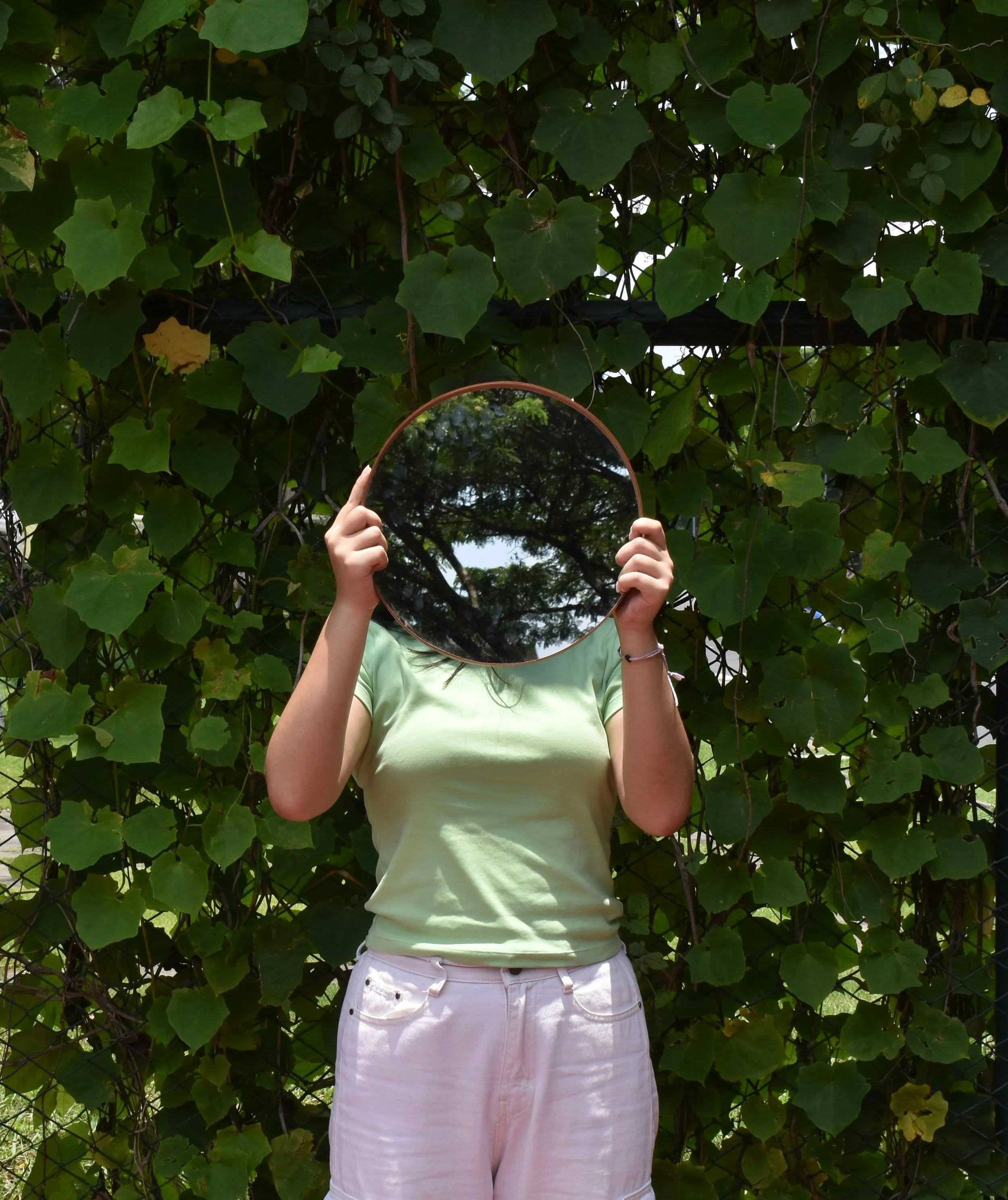 Person standing outdoors, holding a mirror in front of their face, with green leafy vines and foliage behind them.