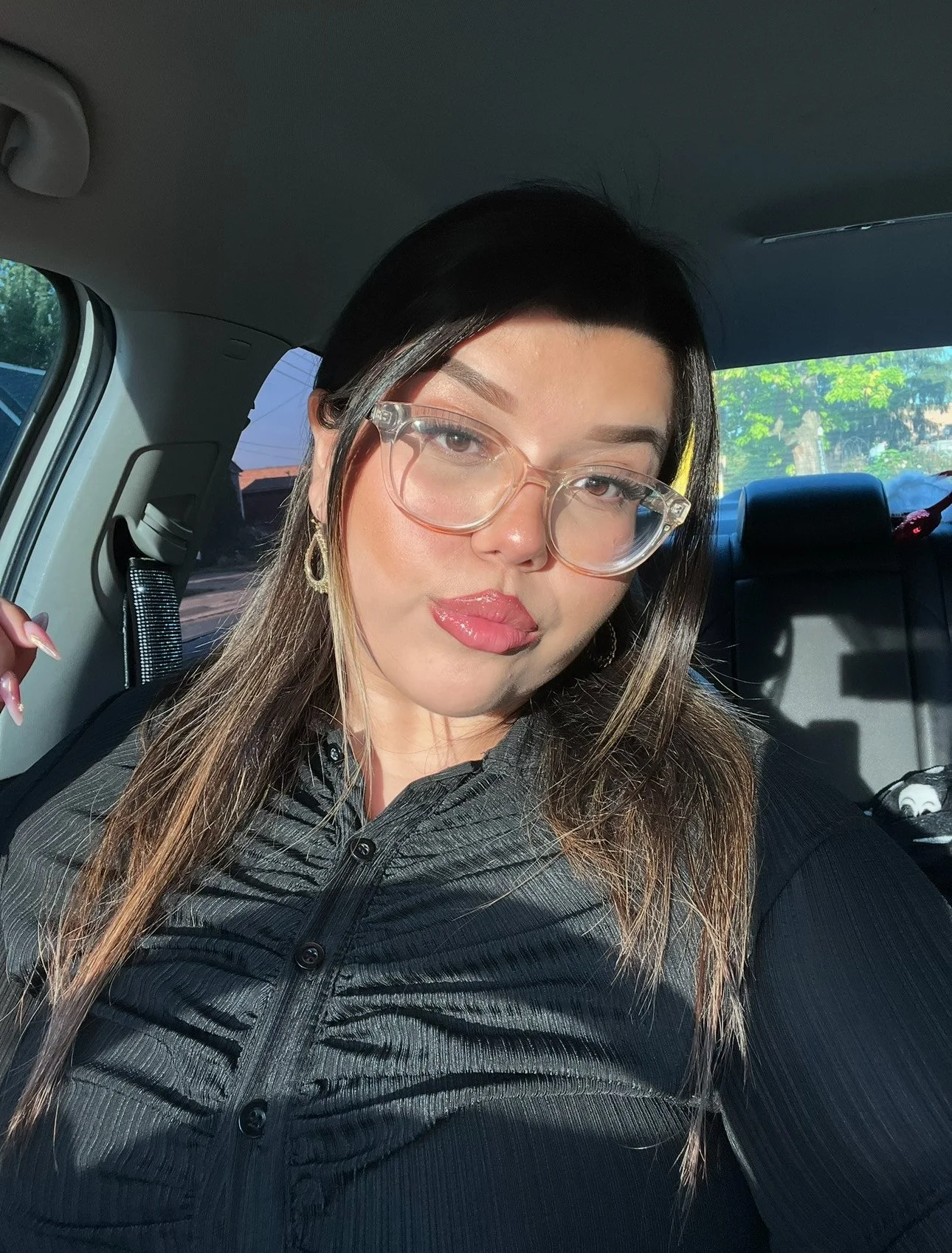 A woman with long brown hair, wearing clear glasses and gold earrings, sitting in the backseat of a car. She is wearing a black ribbed top and has a slight pout on her lips, with sunlight illuminating her face.