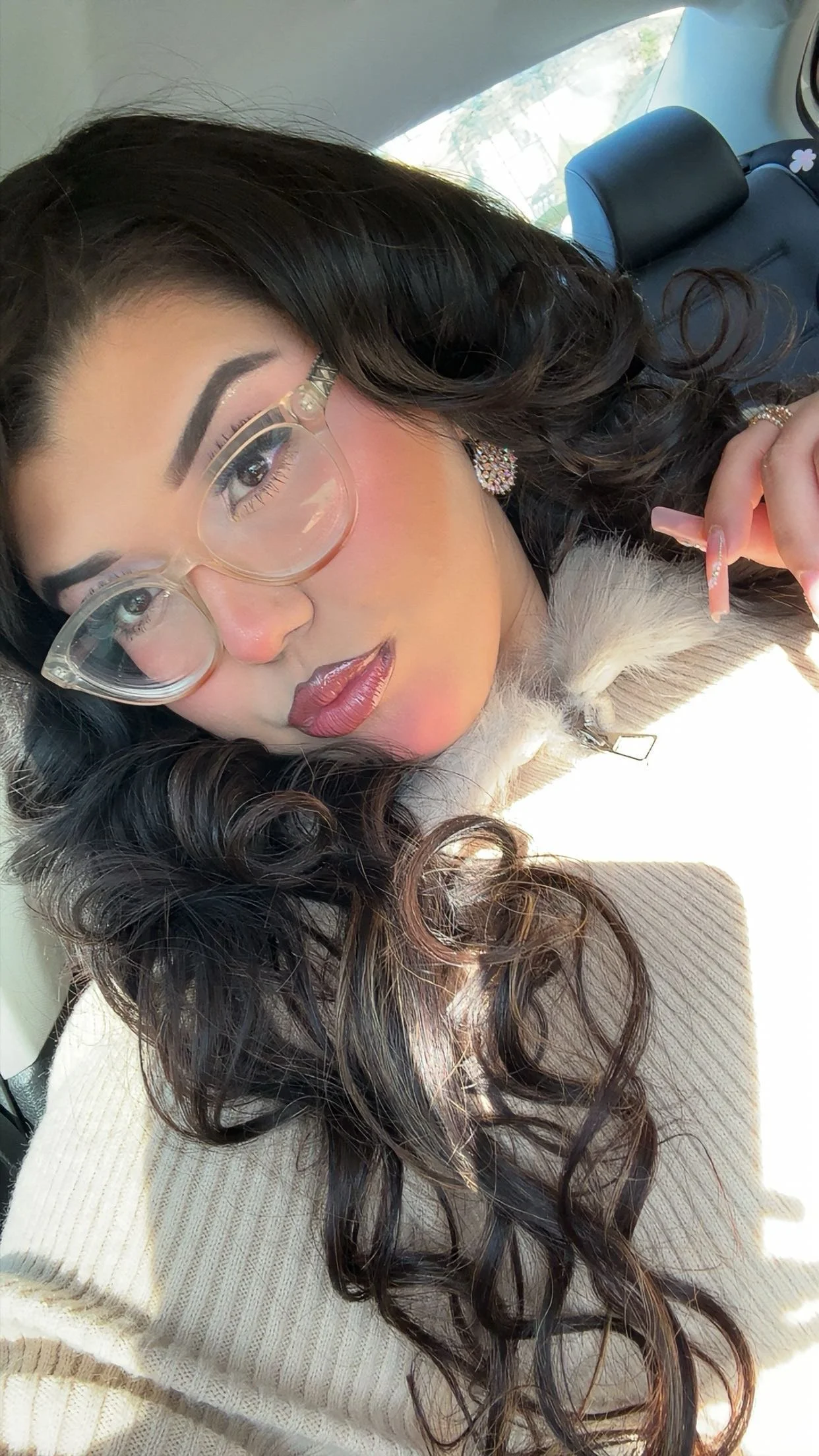 A woman with curly dark hair, wearing glasses with clear frames, earrings, and makeup, sitting in a car with sunlight on her face, holding a cigarette.