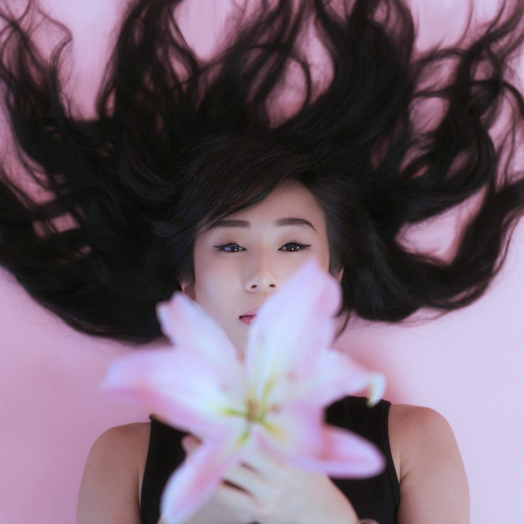 A woman with long black hair lying on a pink background, holding a large pink and white lily flower in front of her face.
