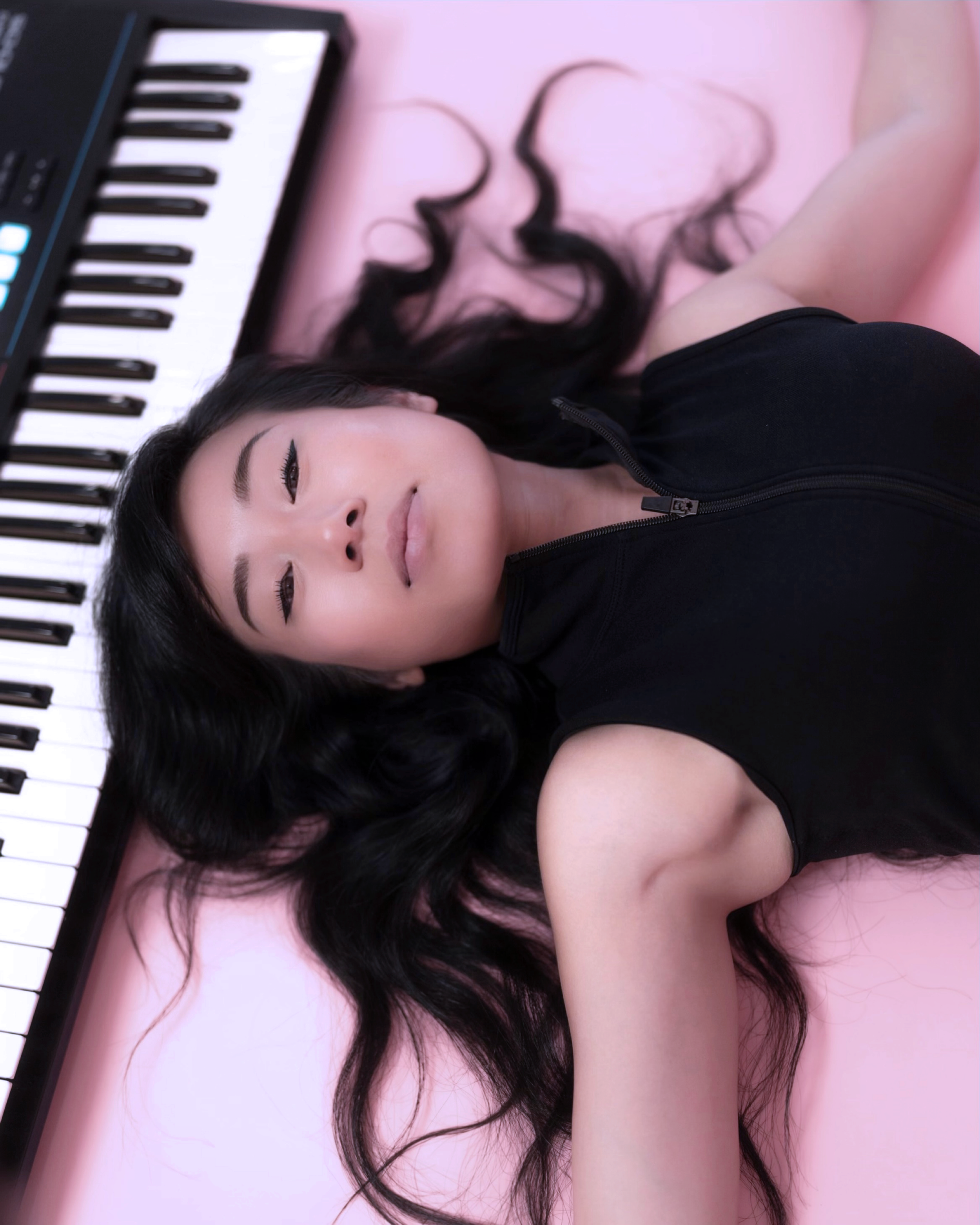 A woman with long black hair lying on a pink surface next to a keyboard synthesizer, wearing a black sleeveless top with a zipper.