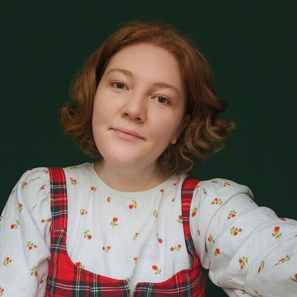 an image of Ella smiling into the camera with her head slightly tilted to the right. She is wearing a white shirt patterened with little red roses, and a red vest over the top. She had bob-length curly red hair.
