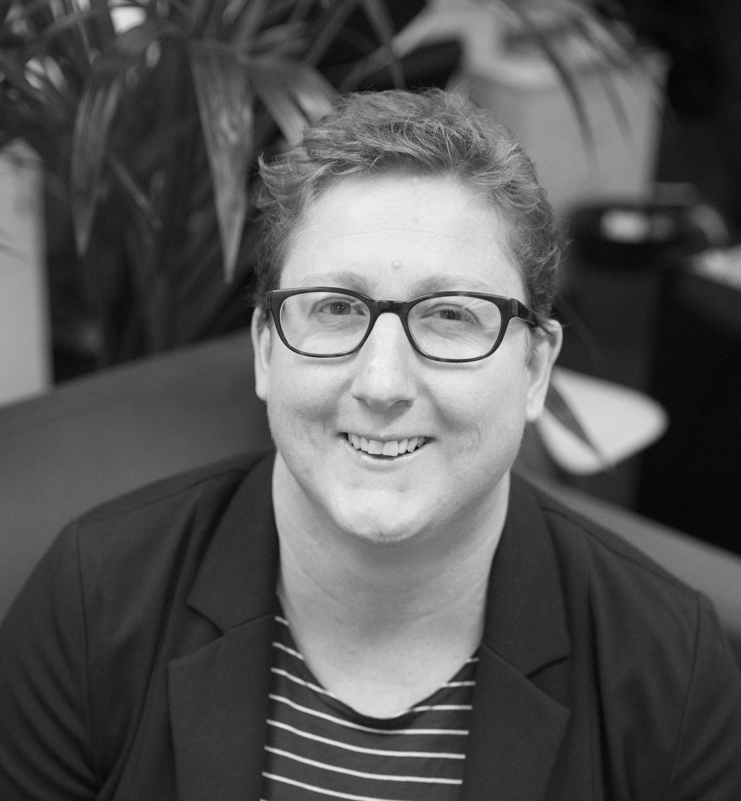 Black and white image of a woman wearing a striped shirt and jacket and glasses against the plant in the background.