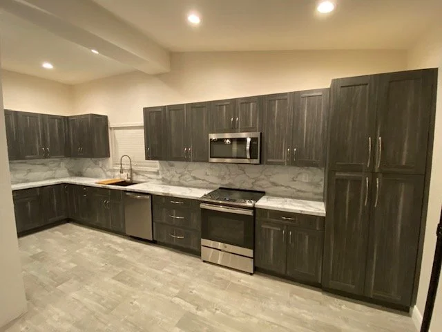 Modern kitchen with dark wood cabinets, marble backsplash, stainless steel appliances, and light-colored flooring.