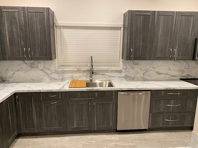 Modern kitchen with gray cabinets, white marble backsplash, stainless steel dishwasher, and a window with blinds above the sink.