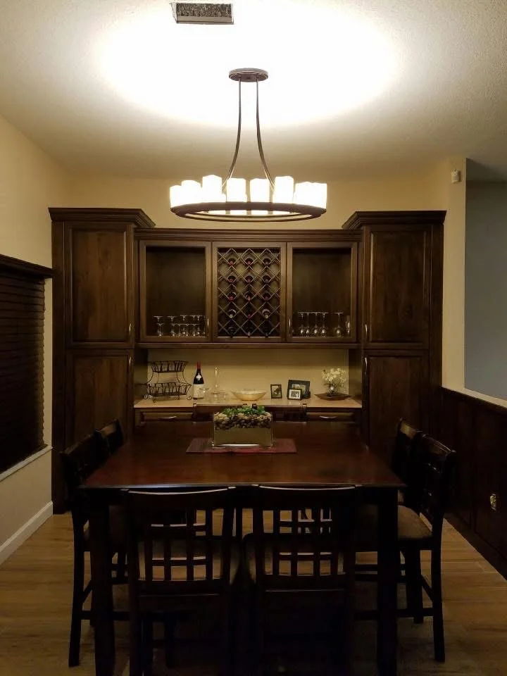 A dining room with a wooden table surrounded by six chairs, a large wooden cabinet with glassware and wine rack, and a chandelier hanging from the ceiling, illuminated by a ceiling light.