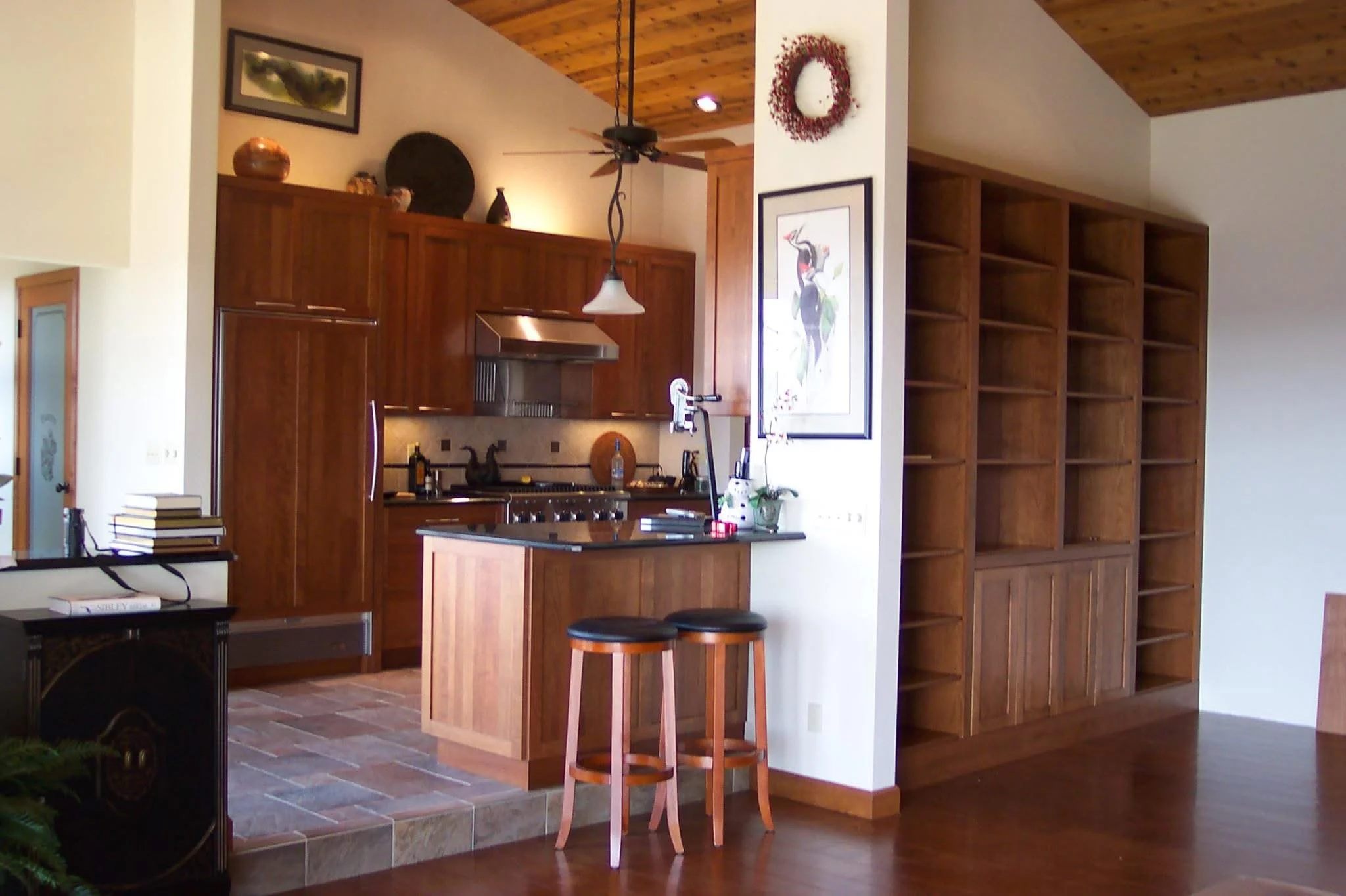 A kitchen with wooden cabinets, a small island with two bar stools, a built-in open wooden shelving unit, and artwork on the wall.