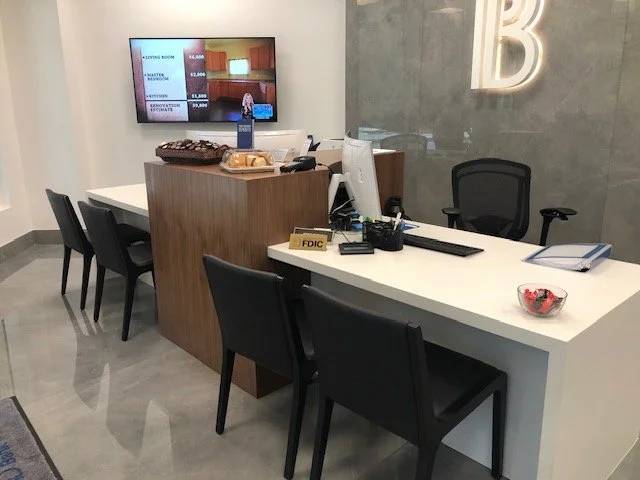 Office reception area with a white desk, black chairs, a computer, and a small bowl of candies, with a television mounted on the wall displaying advertisement or information.