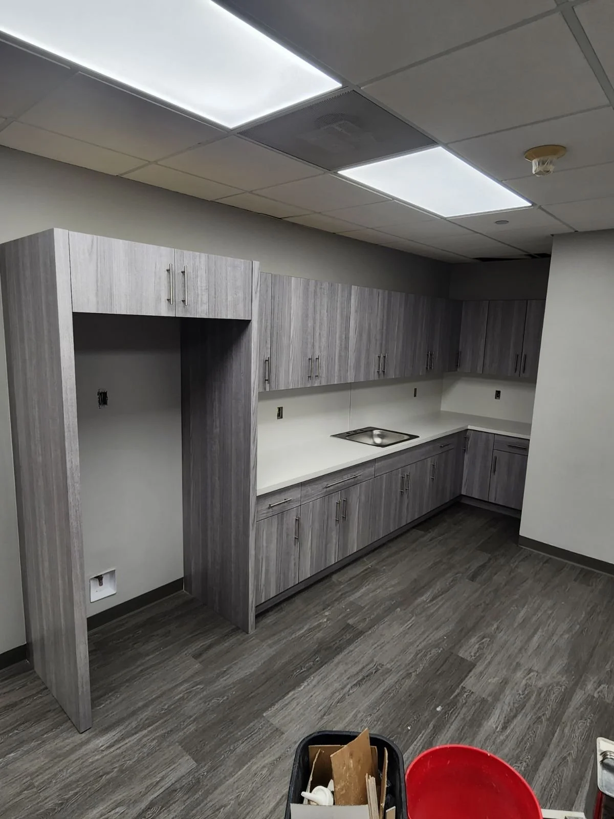 Empty kitchenette with gray cabinets and white countertops, ceiling lights, and a small sink, with construction supplies in the foreground.