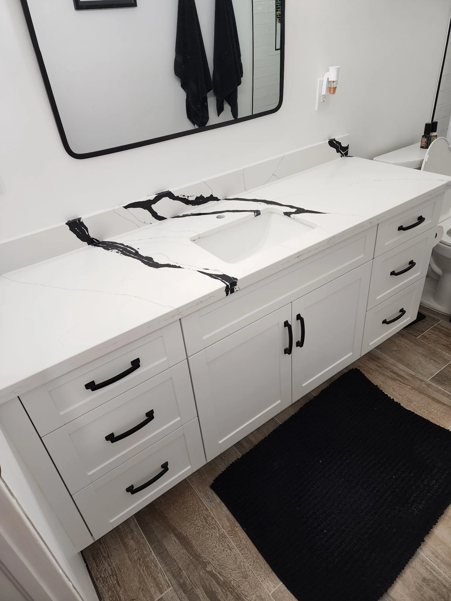 Bathroom vanity with white marble countertop and black veining, mirror above, black handles on white cabinets, black rug on brown tile floor, and black towels reflected in mirror.