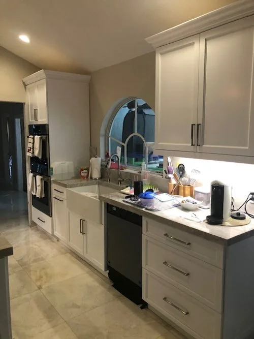 Modern kitchen with white cabinets, marble floor, and arched window above the double sink, cluttered with dish soap, utensils, and appliances.