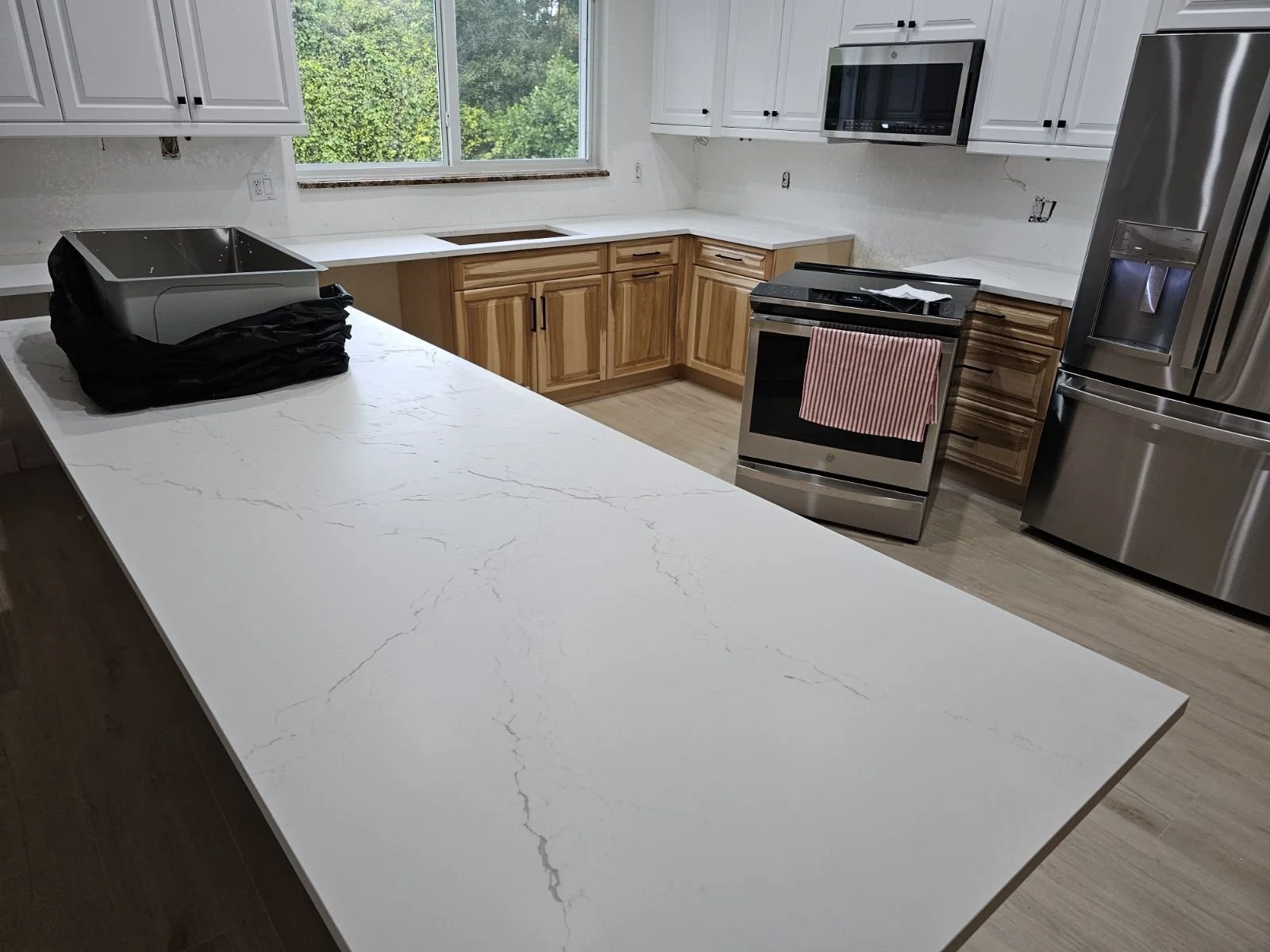 Modern kitchen with white countertops, wooden cabinets, stainless steel appliances including a refrigerator, oven, and microwave, and a large window overlooking green foliage.