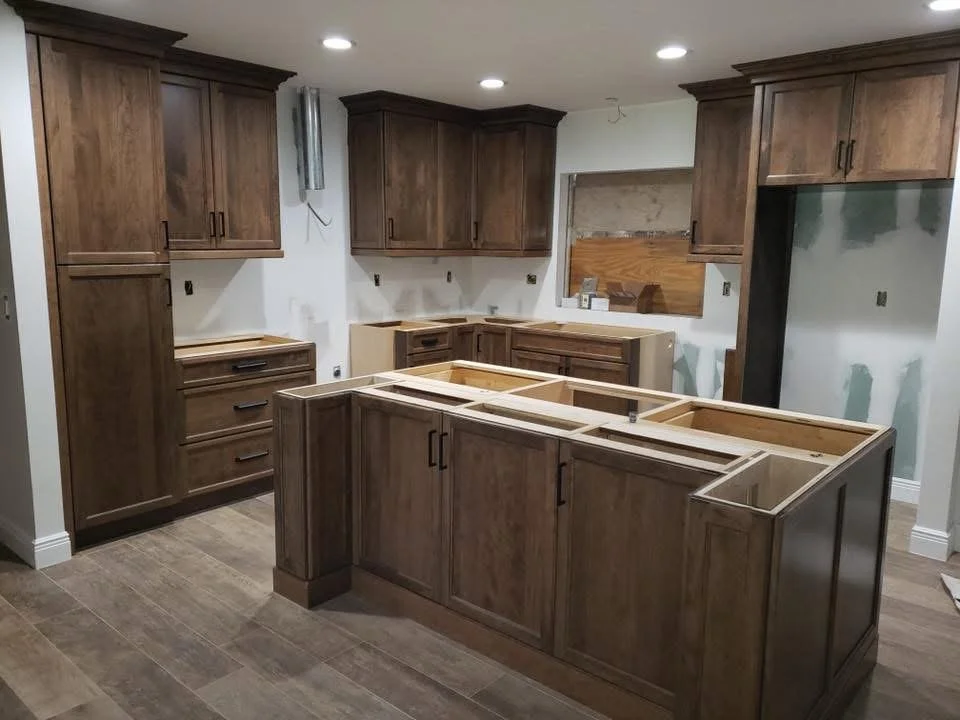 Kitchen cabinets being installed in a new kitchen with brown wood cabinetry, some with drawers, and a central island frame without countertop, in a room with recessed lighting and wood-look floor tiles.