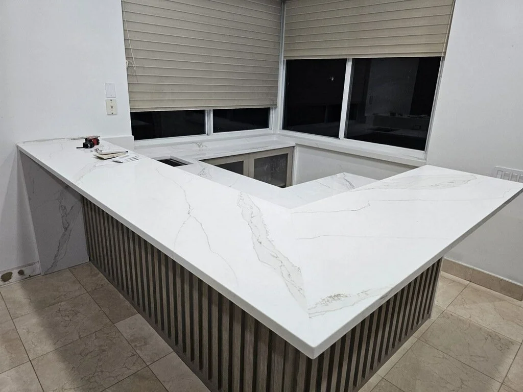 A white marble kitchen island with a rectangular cutout, featuring vertical wooden slats on the sides, in a room with large windows covered by horizontal beige blinds.