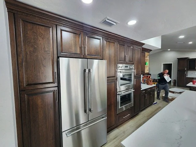 Modern kitchen with dark wood cabinets, stainless steel refrigerator, and built-in oven. A person is standing near the countertop.