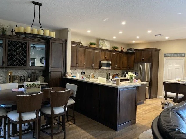 Kitchen with dark wood cabinets, stainless steel refrigerator, microwave, and white countertops; dining area with chairs; small dog near a couch.