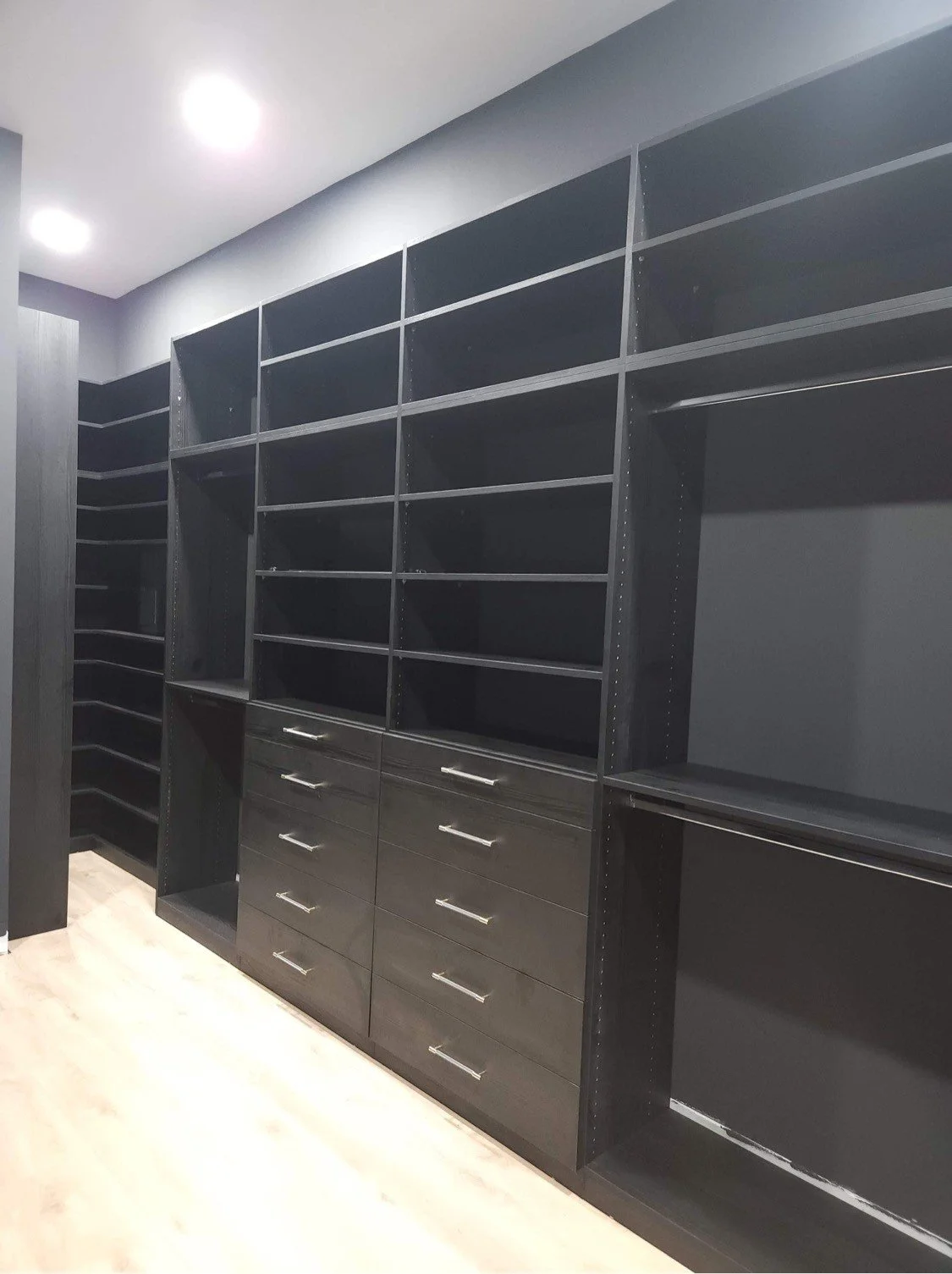 A modern, empty black closet with multiple shelves, drawers, and hanging rods, in a room with light wood flooring and ceiling lights.